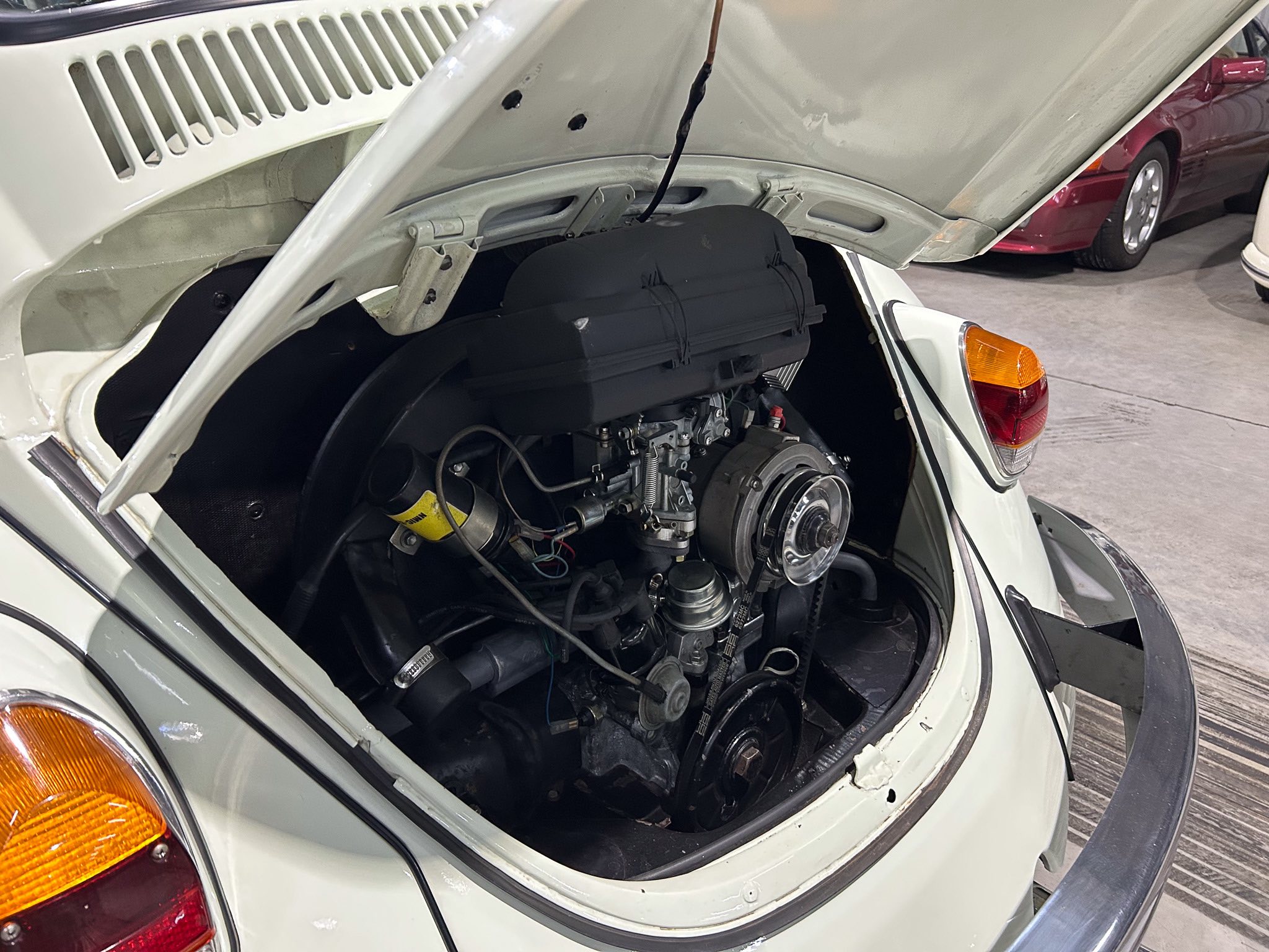 Rear-hinged trunk open on a white vintage car, exposing the engine and various hoses and belts in the compartment