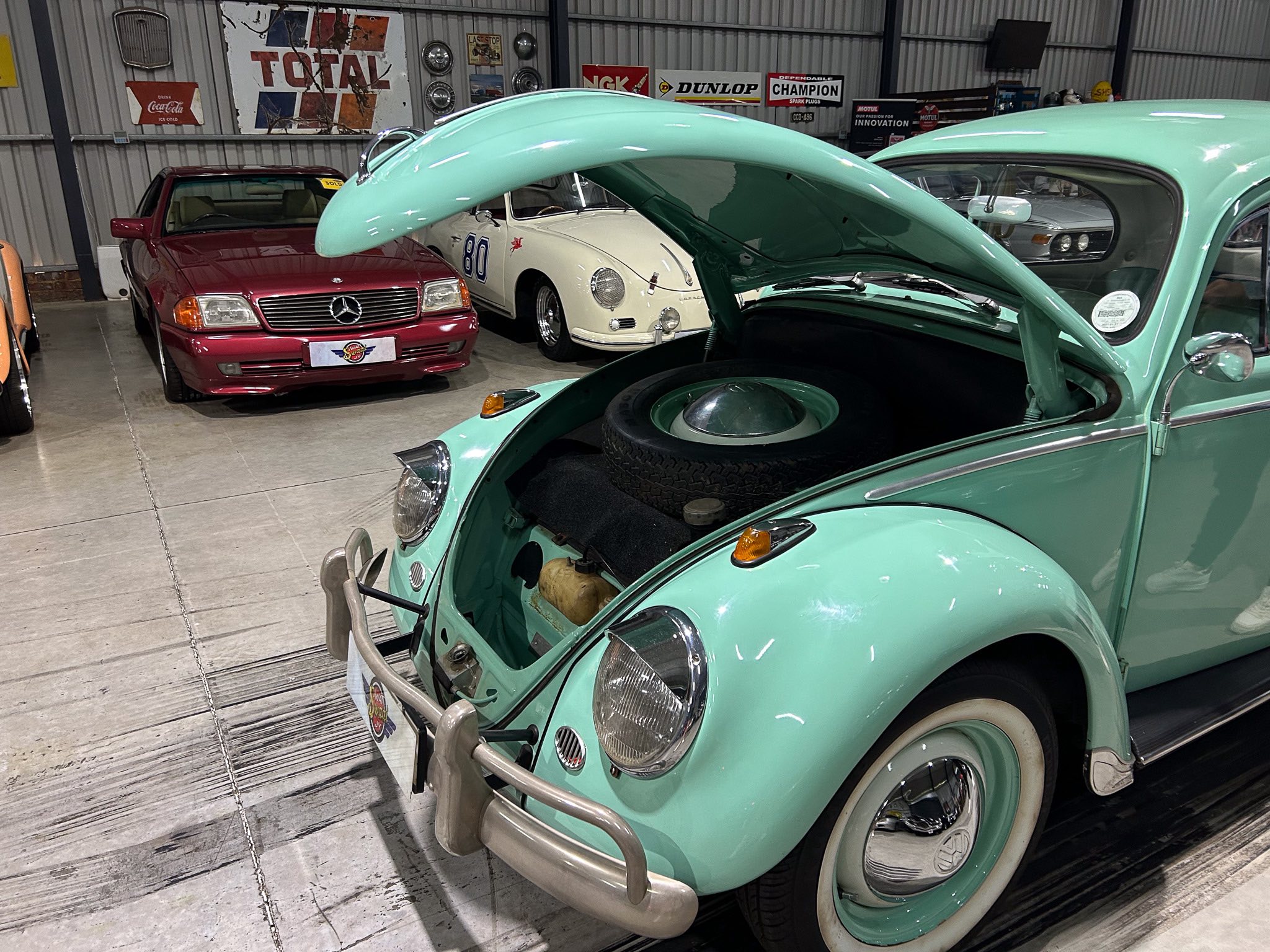 Mint green vintage VW Beetle with hood open in a garage showroom, exposing spare tire and engine area; other classic cars and retro signage in background.