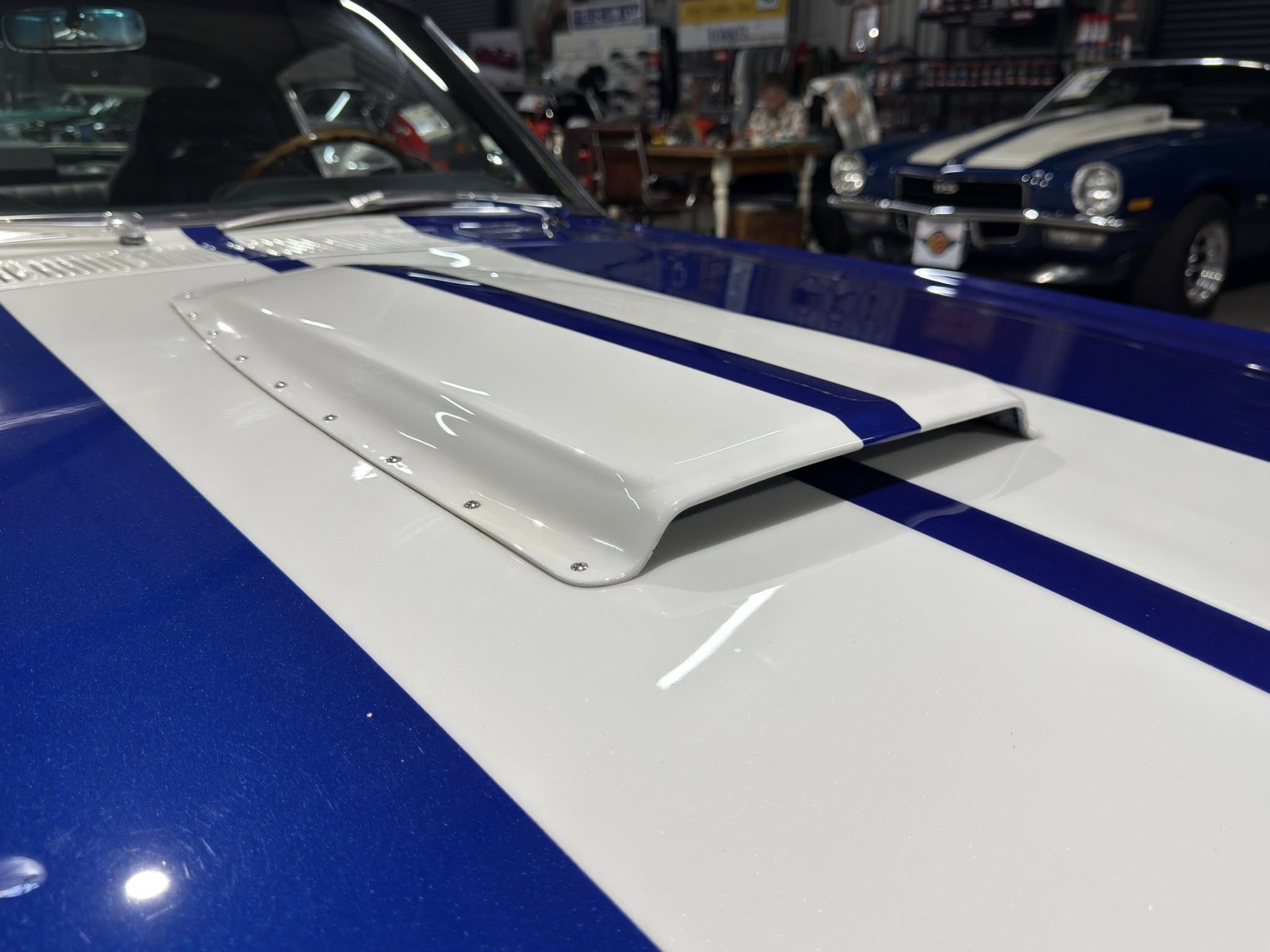 Close-up of a blue car hood with a white racing stripe in a showroom setting.