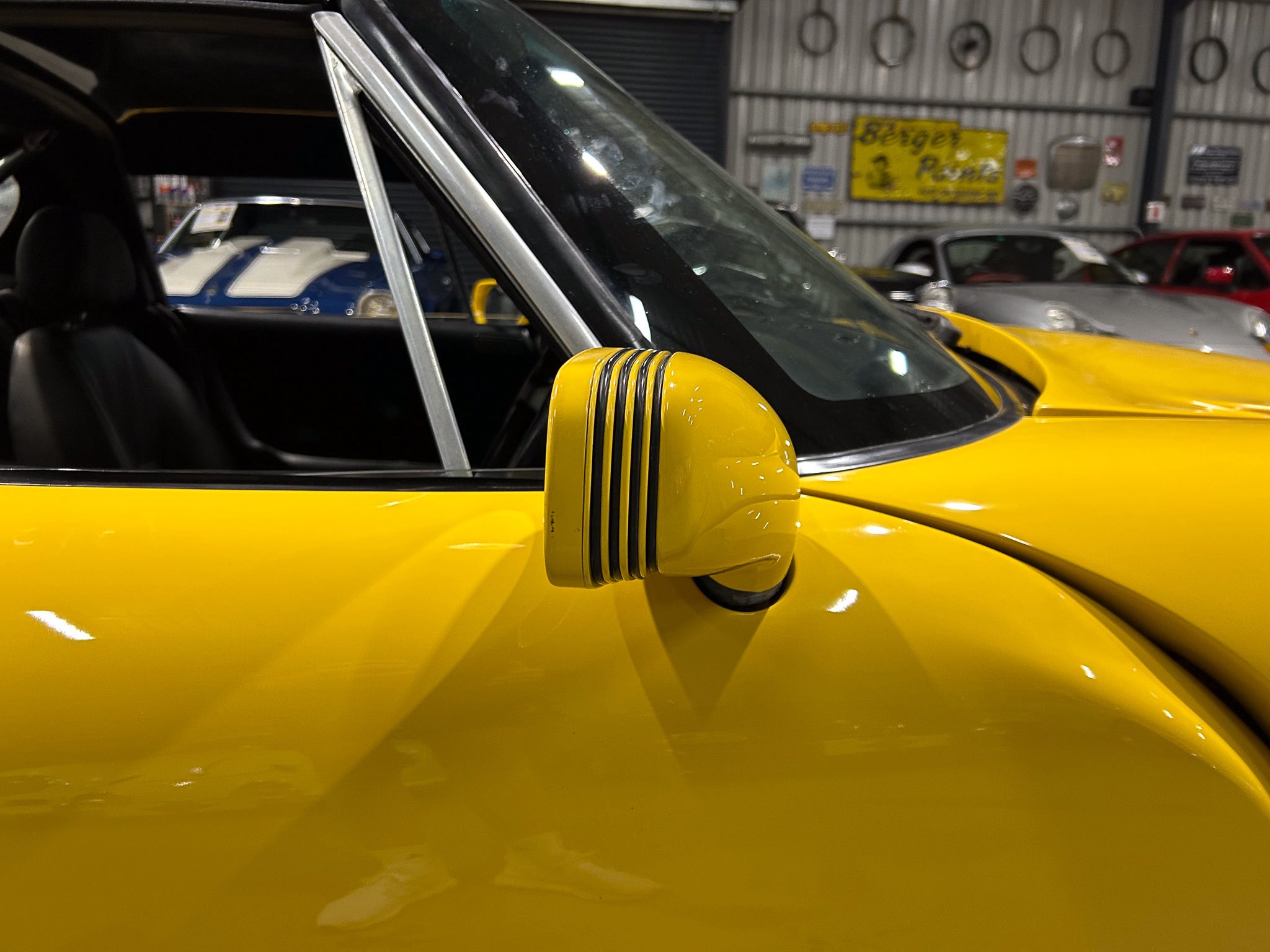 Close-up of a yellow car door with chrome window frame and vent details, showroom background with other cars visible behind it.
