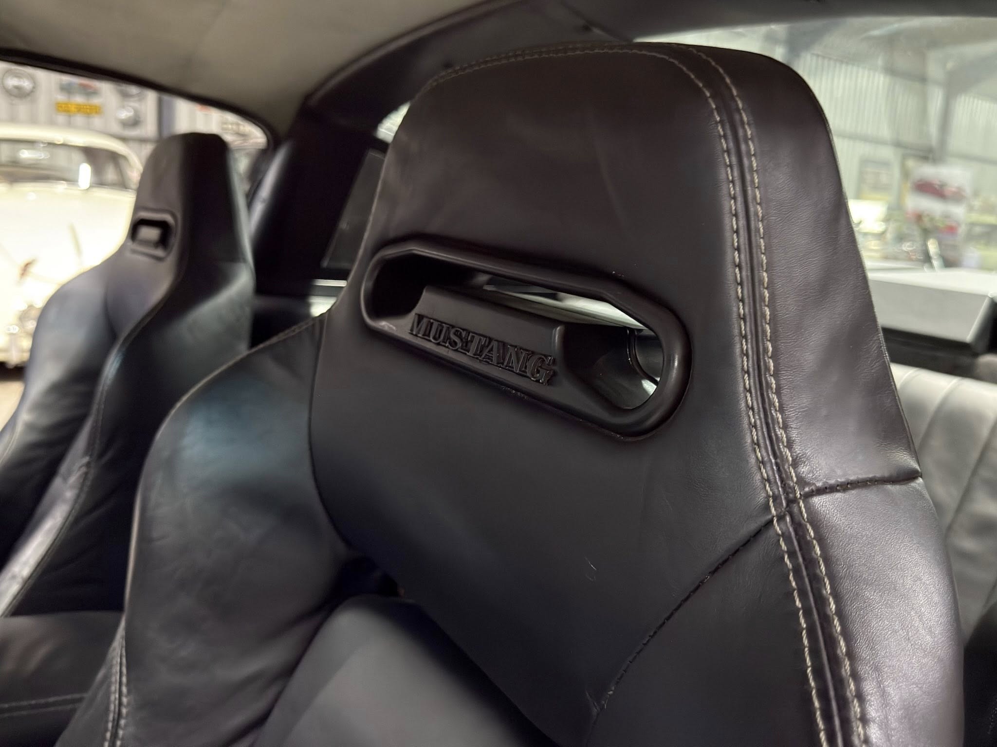 Black leather car seat with white contrast stitching and 'MUSTANG' branding visible in a vehicle interior.
