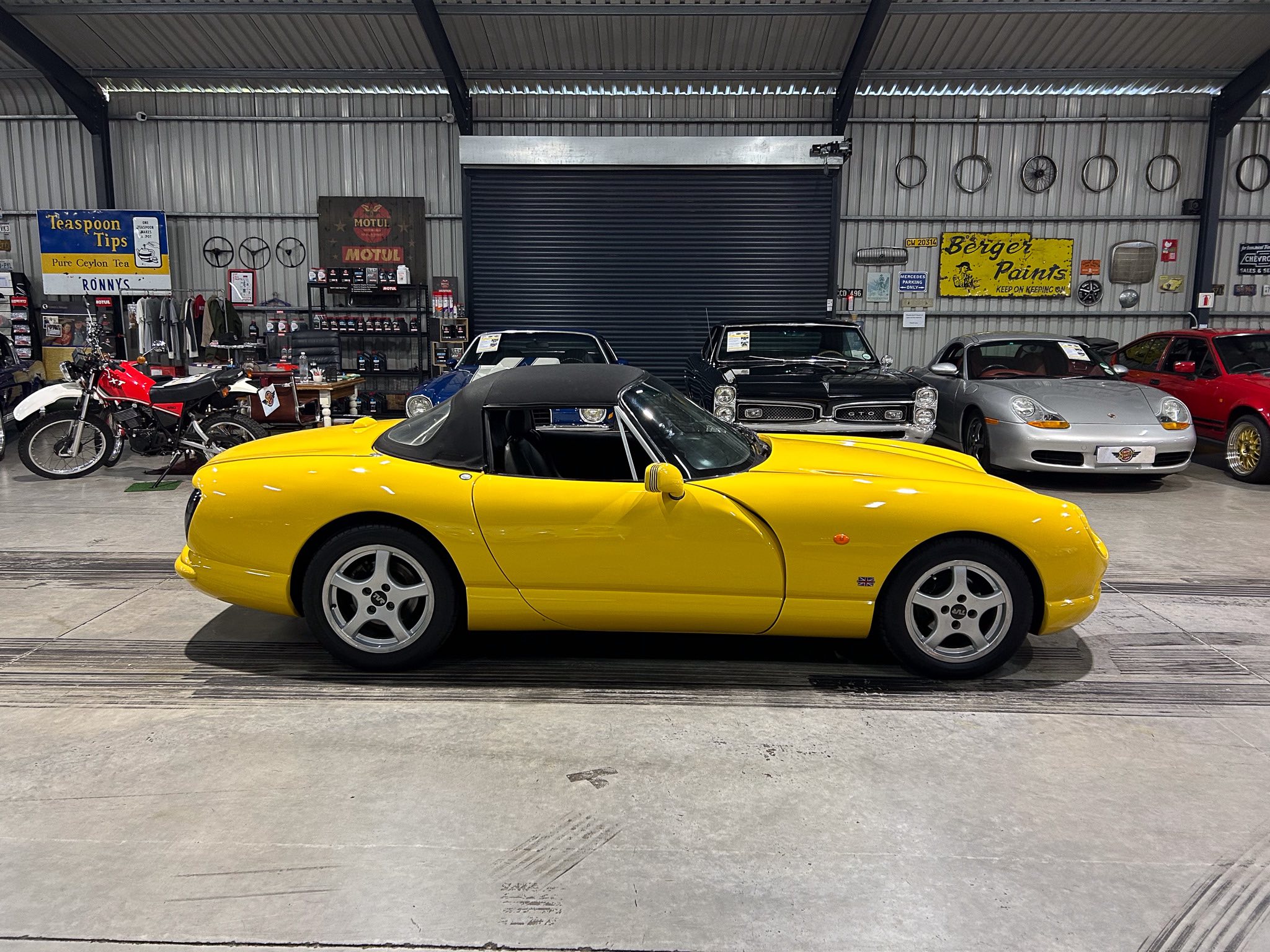 Yellow convertible sports car in a showroom/garage with vintage signs and other vehicles in the background.