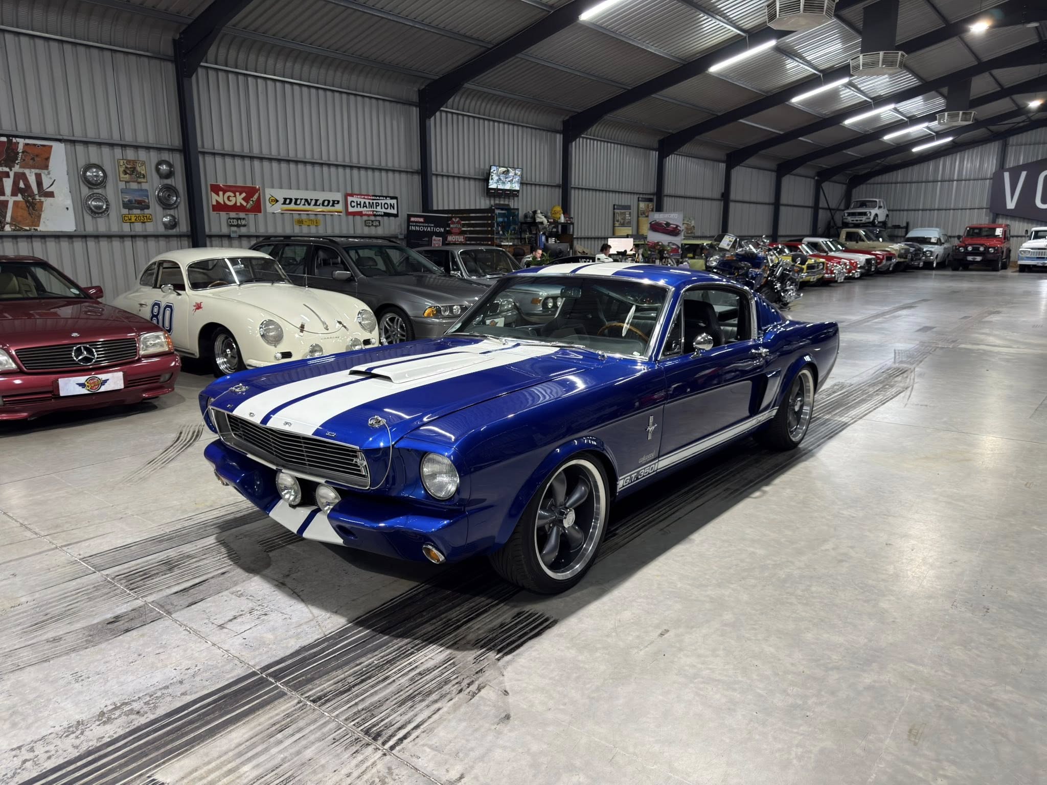 Blue Ford Mustang with white racing stripes in the foreground of a vintage car showroom filled with classic cars in a large warehouse-like space.