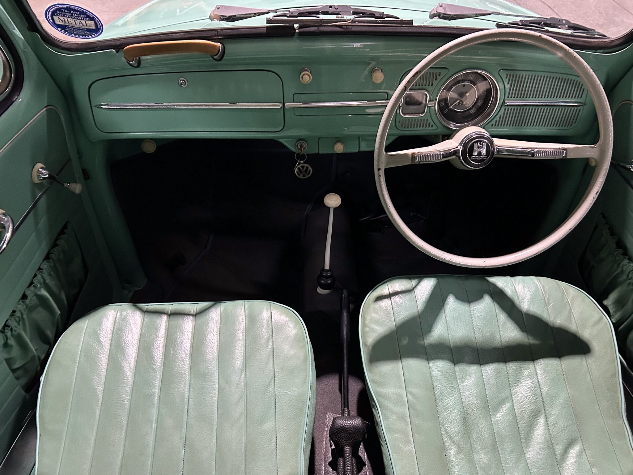 Interior of a vintage green car: two green seats, large white steering wheel with emblem, and a teal dashboard with a round speedometer cluster on the right side