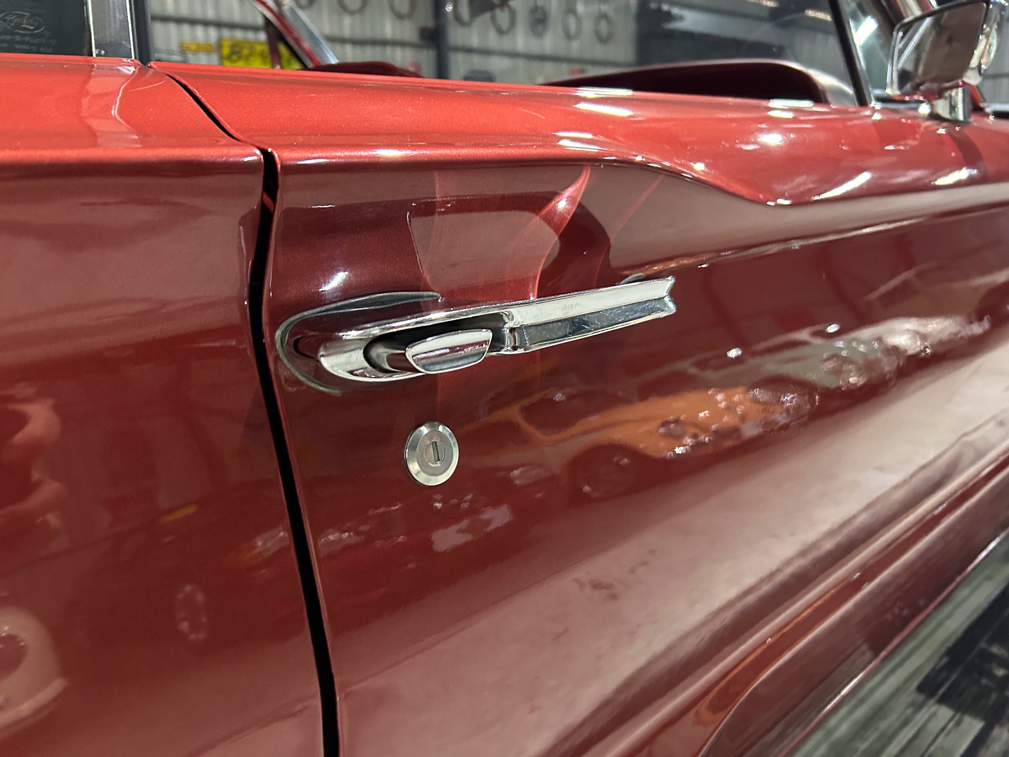 Close-up of a glossy red vintage car door with chrome handle and keyhole, showing a slight dent and reflections in the paint.