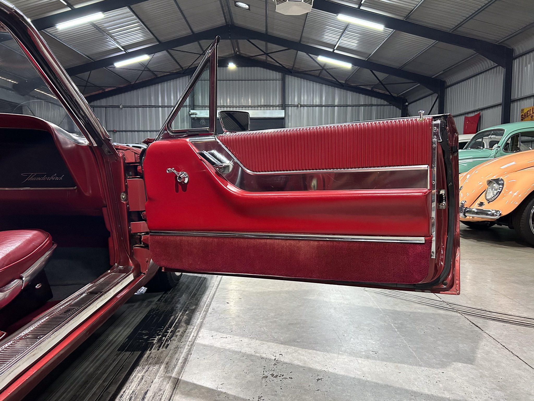 Red vintage car interior with door fully open, showing red pleated door panel and chrome trim in a showroom of classic cars.