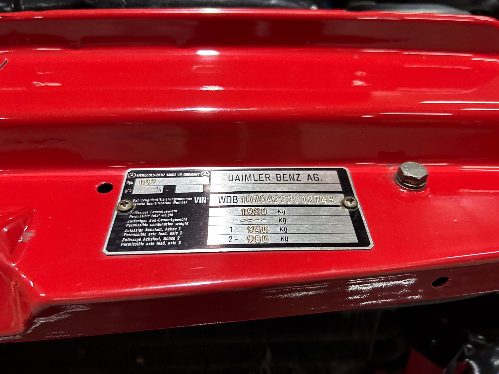 Close-up of a metal vehicle identification plate on a red car, showing VIN and weight information under a red-painted surface.