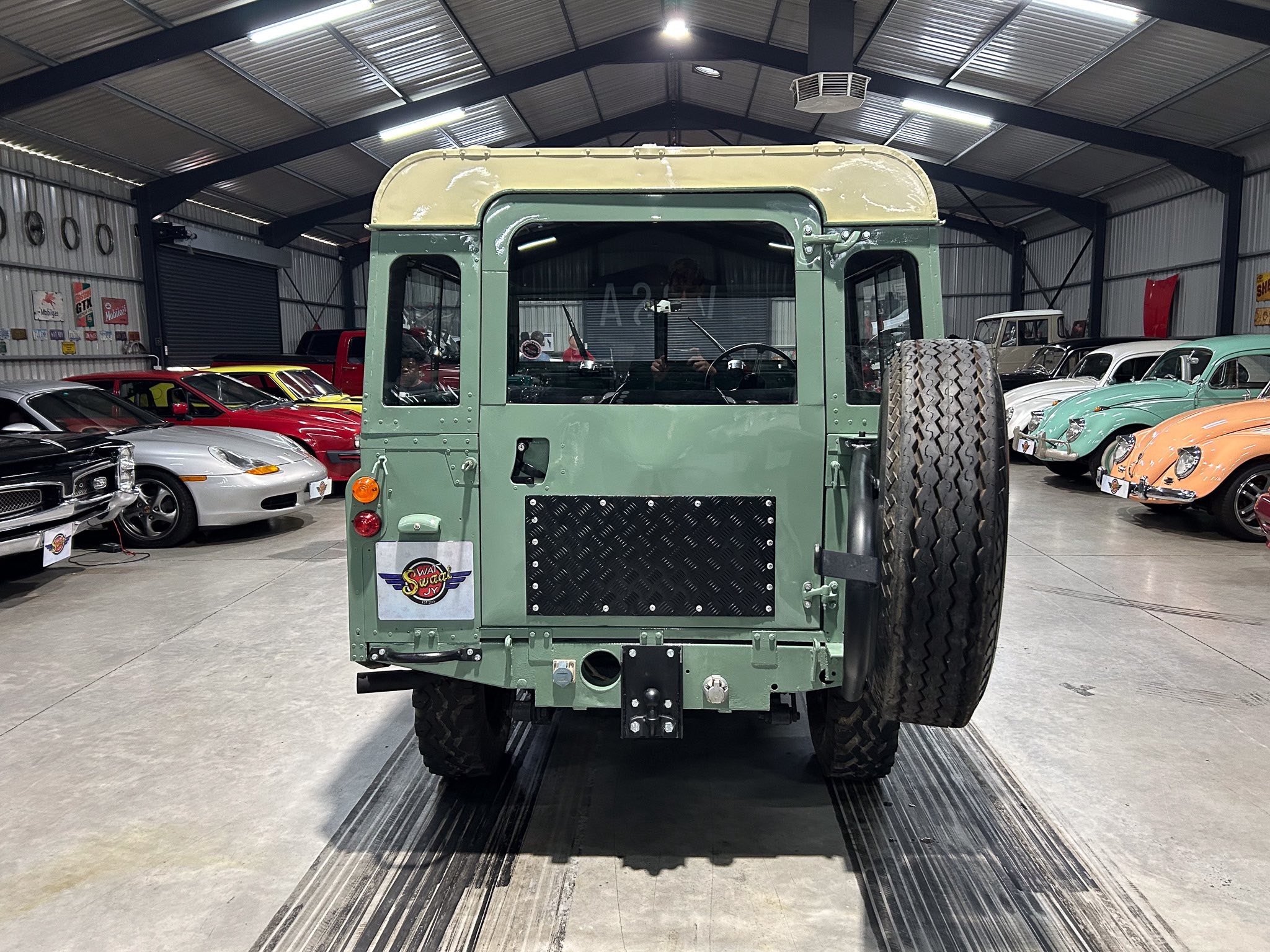 Rear view of a green vintage military jeep inside a showroom with classic cars lining both sides.