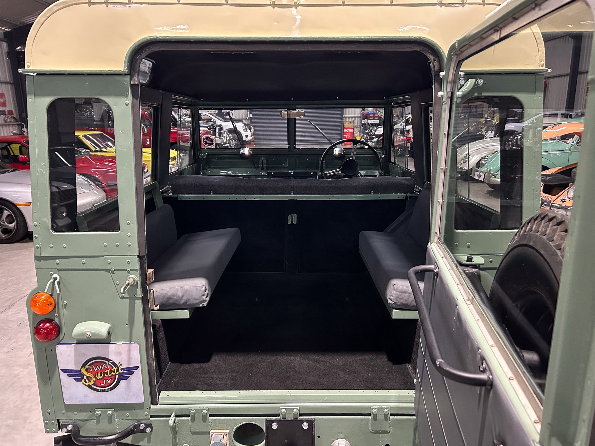 Inside view of a vintage green military truck with an open rear cargo area and side benches, steering wheel visible, showroom of classic cars in the background