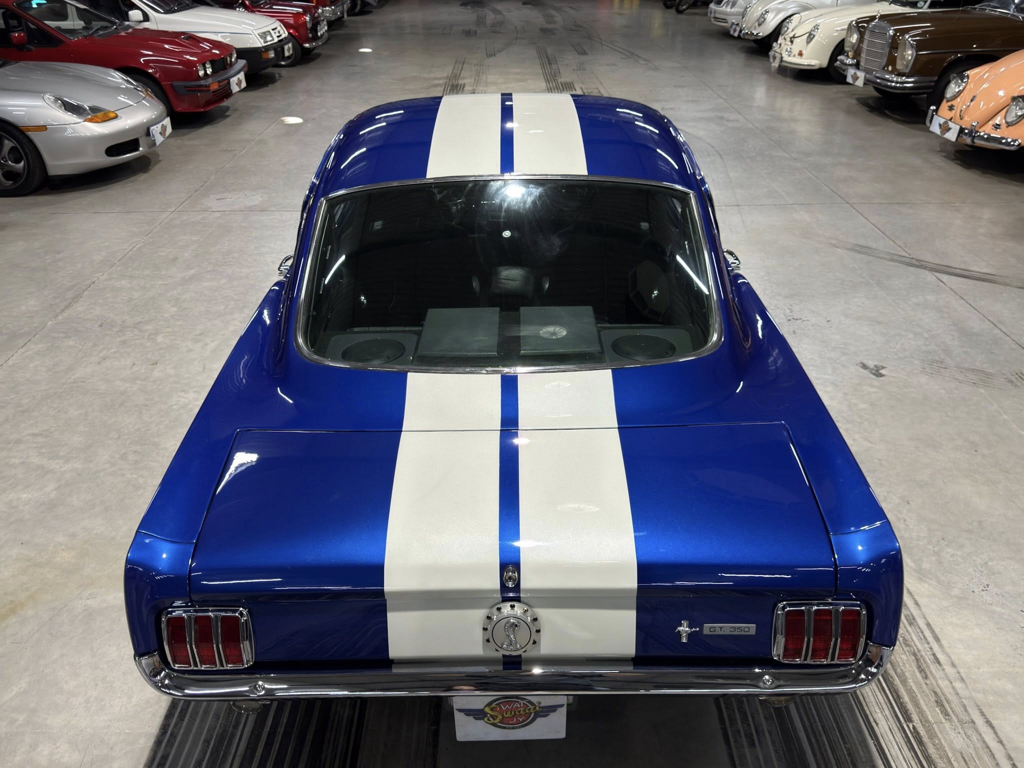 Rear view of a blue Ford Mustang GT350 with a white racing stripe, parked in a showroom among classic cars.