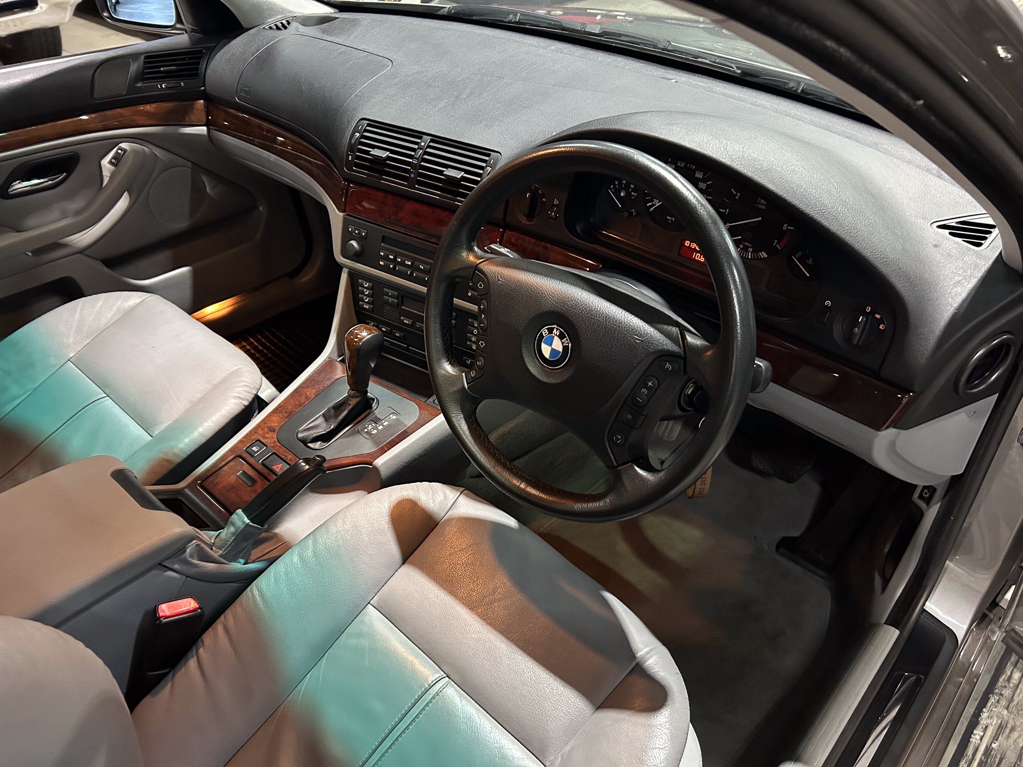 BMW car interior showing the driver's side: steering wheel with logo, dashboard gauges, center console with wood trim and climate controls, and tan leather seats