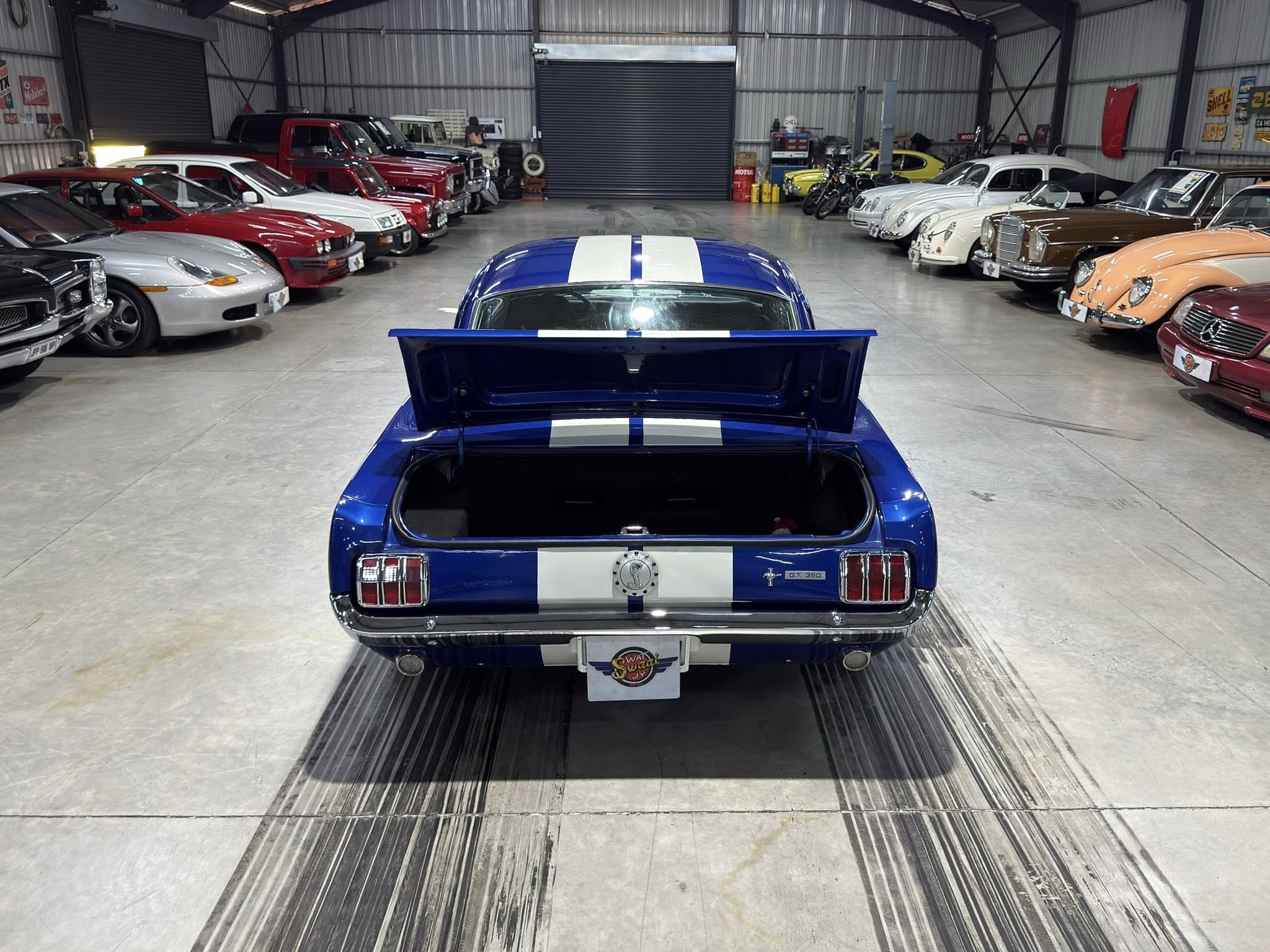 Blue Mustang with white racing stripes, trunk open, seen from the rear in a vintage car showroom lined with classic cars on both sides.