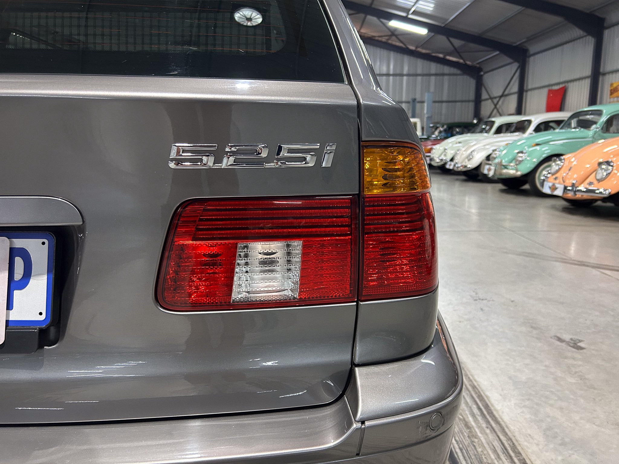 Rear view of a gray car showing the 525i badge and taillight, in a showroom with vintage cars in the background.