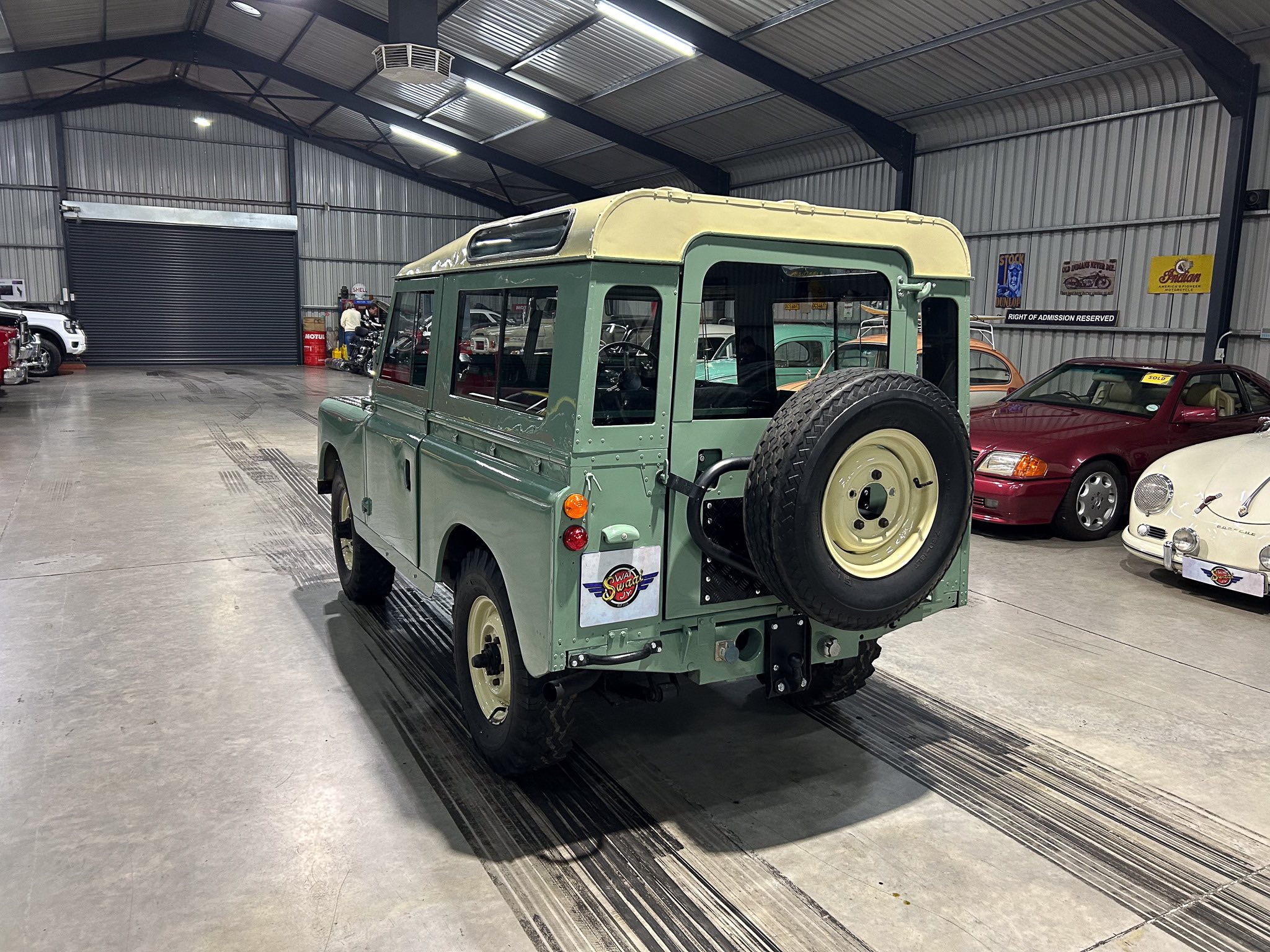 Green vintage off-road vehicle with beige roof inside a garage or showroom, spare tire mounted on the rear door, surrounding classic cars and retro posters on the wall
