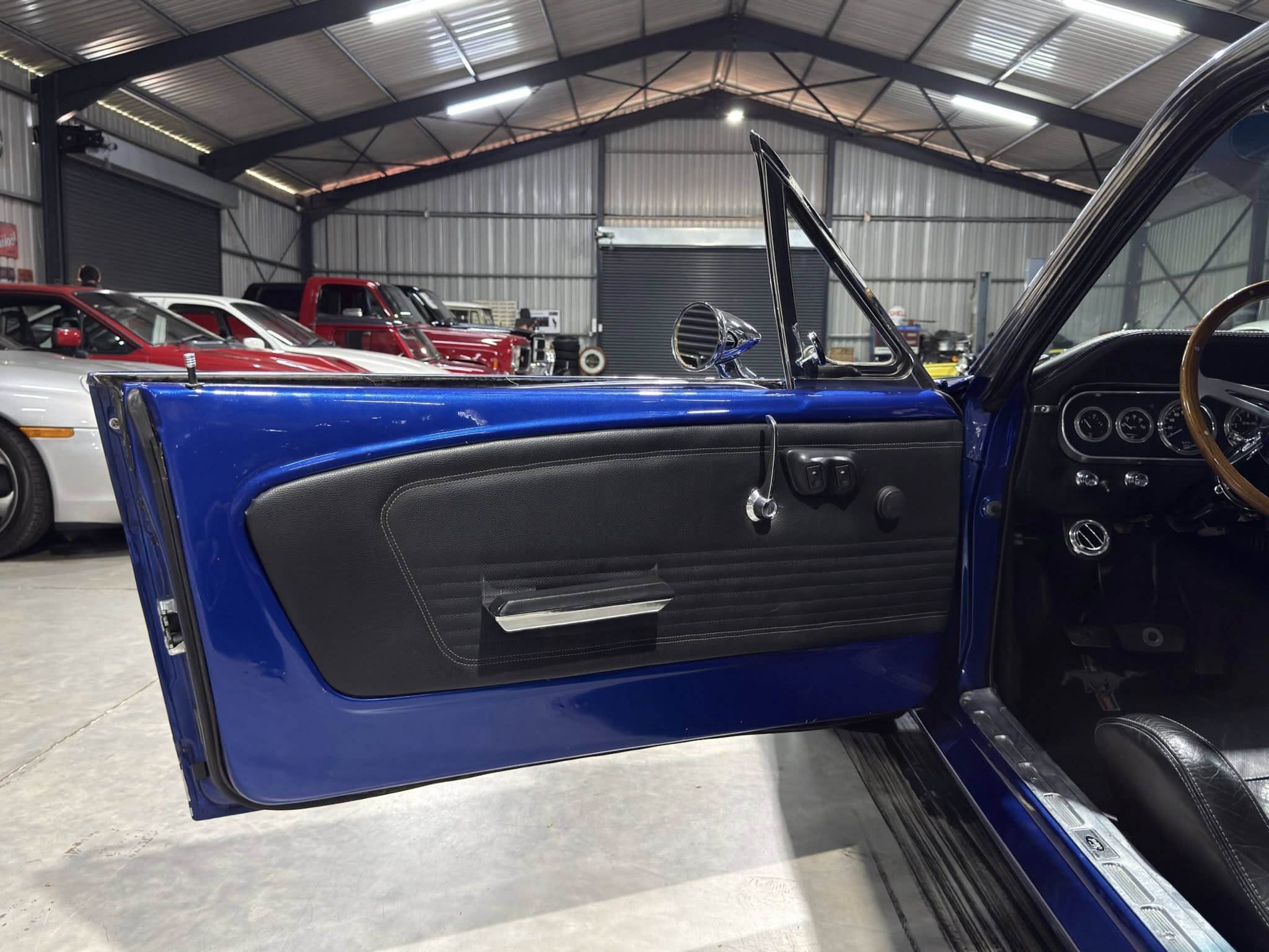 Open blue car door in a showroom/garage, showing the black interior and wood steering wheel with chrome dashboard gauges in the foreground.