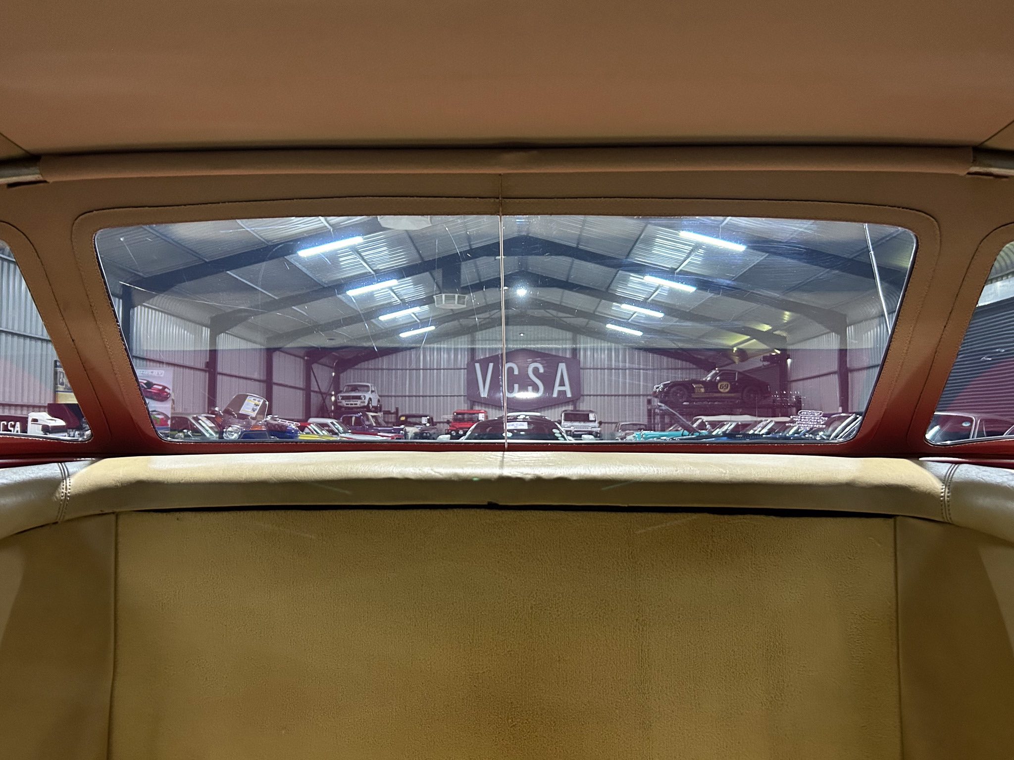 Back-seat view through a tan car rear window onto a showroom filled with vintage cars and a VCSA sign.