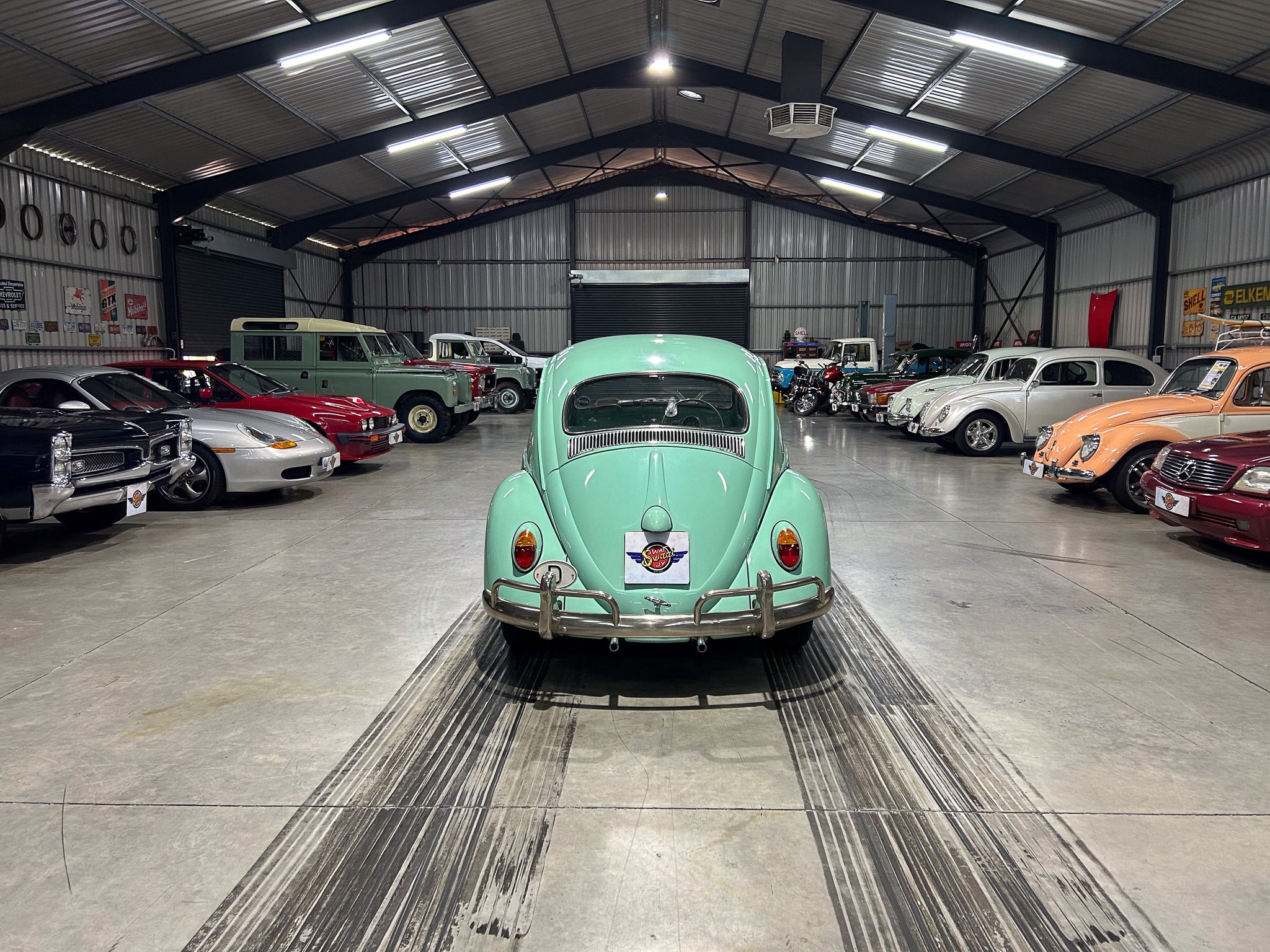 Mint-green vintage car (Beetle) centered in a large indoor showroom, surrounded by a line-up of classic cars.
