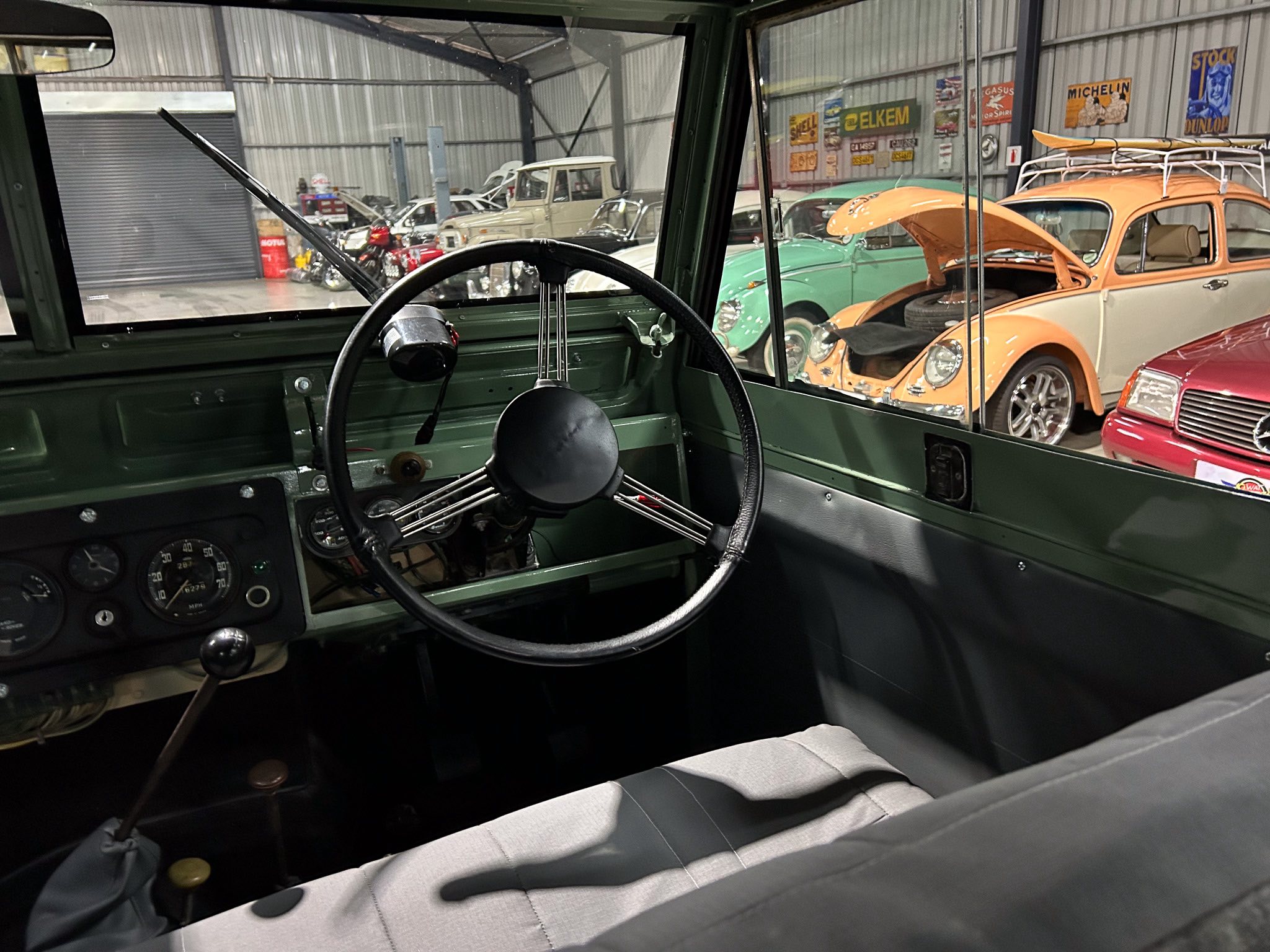 Front-seat view of a classic car interior with a large black steering wheel and vintage gauges; a showroom of retro cars is visible through the windshield.