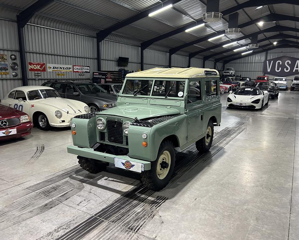 Vintage green Land Rover in a classic car showroom with other antique cars around it indoors at a museum-like space.