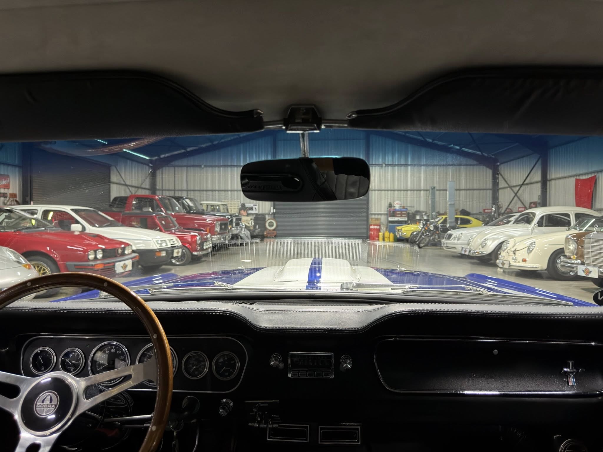 View from the driver’s seat of a vintage car showroom, dashboard instruments visible with many classic cars outside through the windshield.
