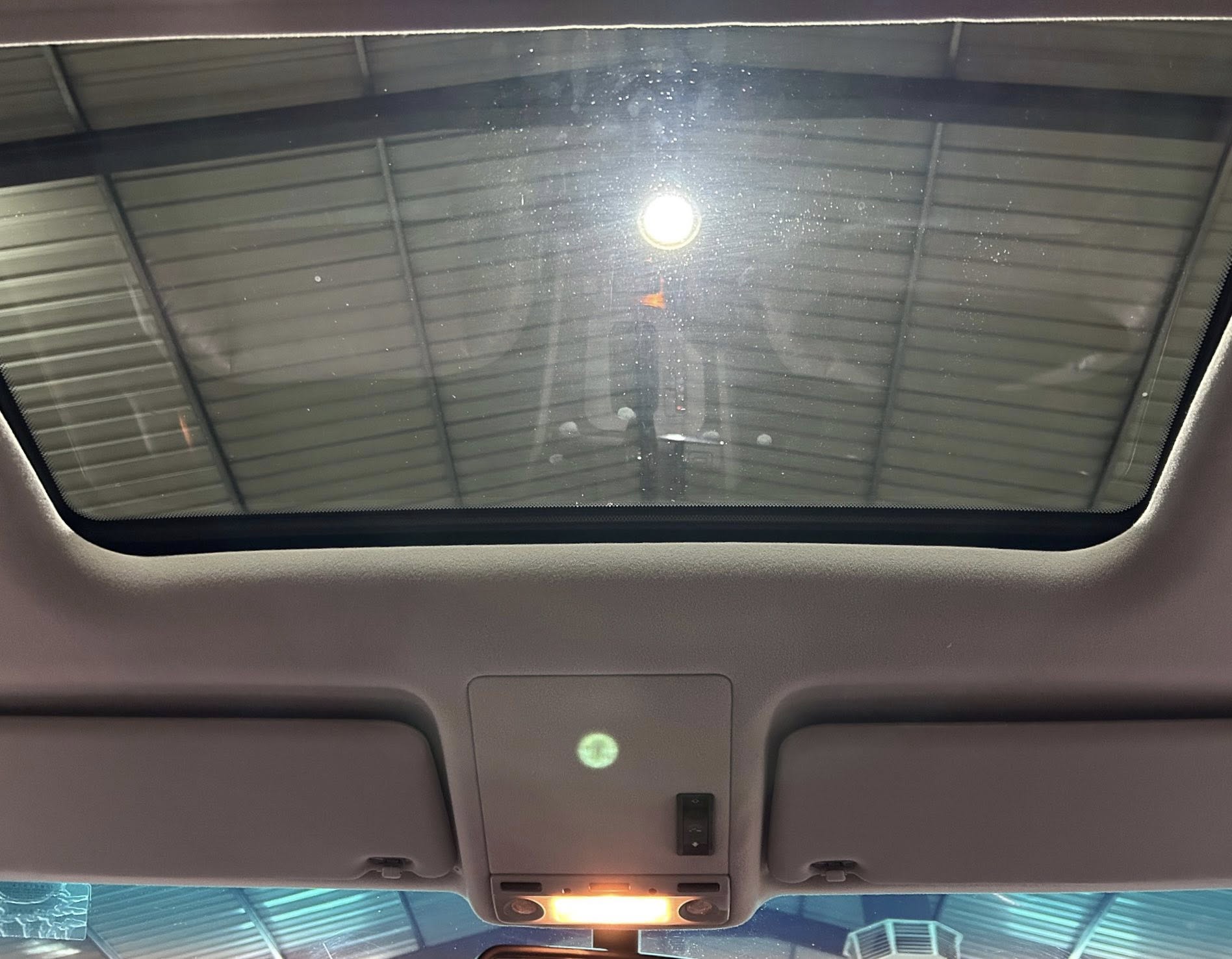 Interior of a car ceiling with a large sunroof, overhead console, and lit cabin lights; garage structure visible through the glass