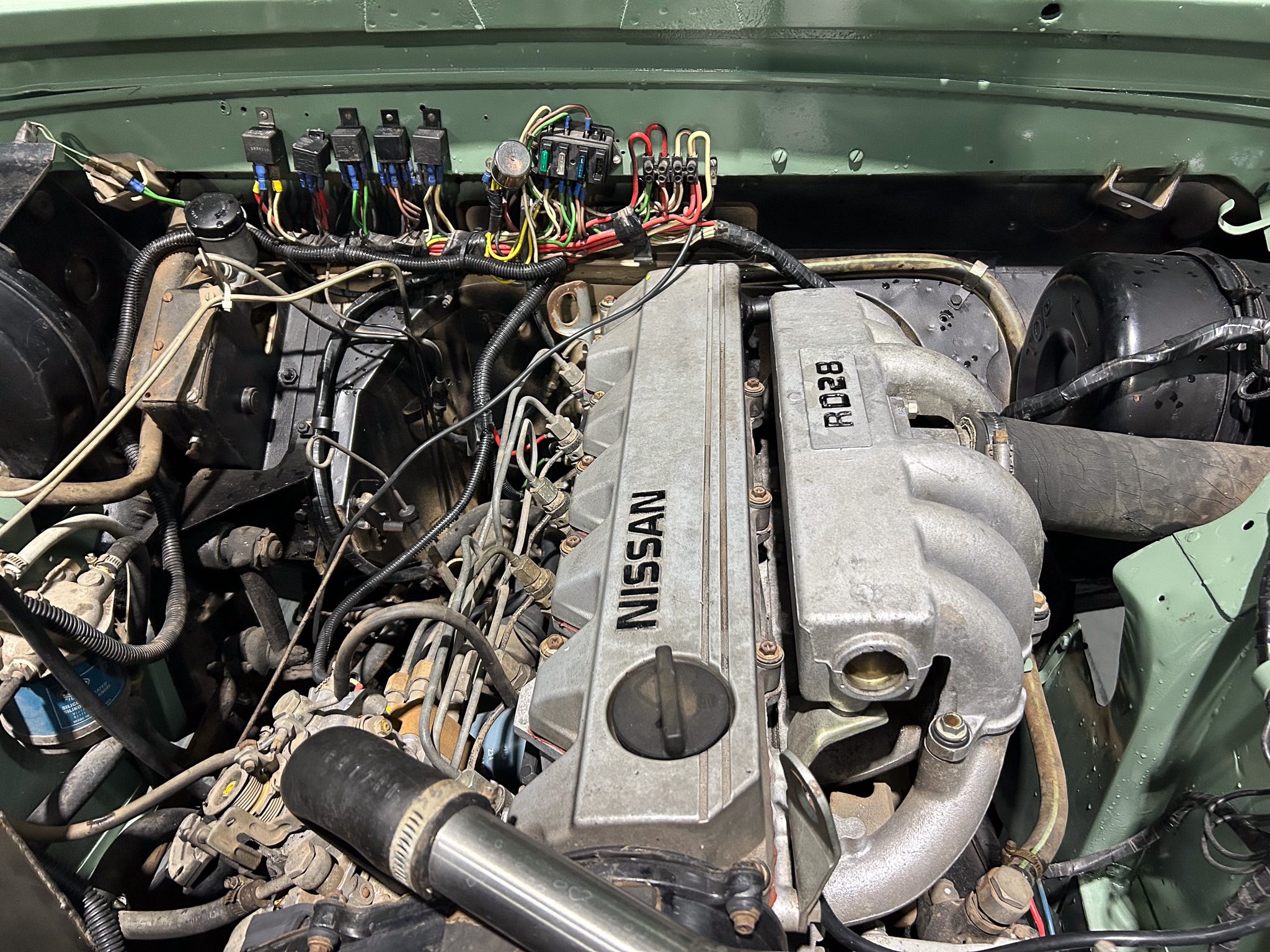 Engine bay of a Nissan showing a messy wiring harness and electrical connectors along a green firewall surface.