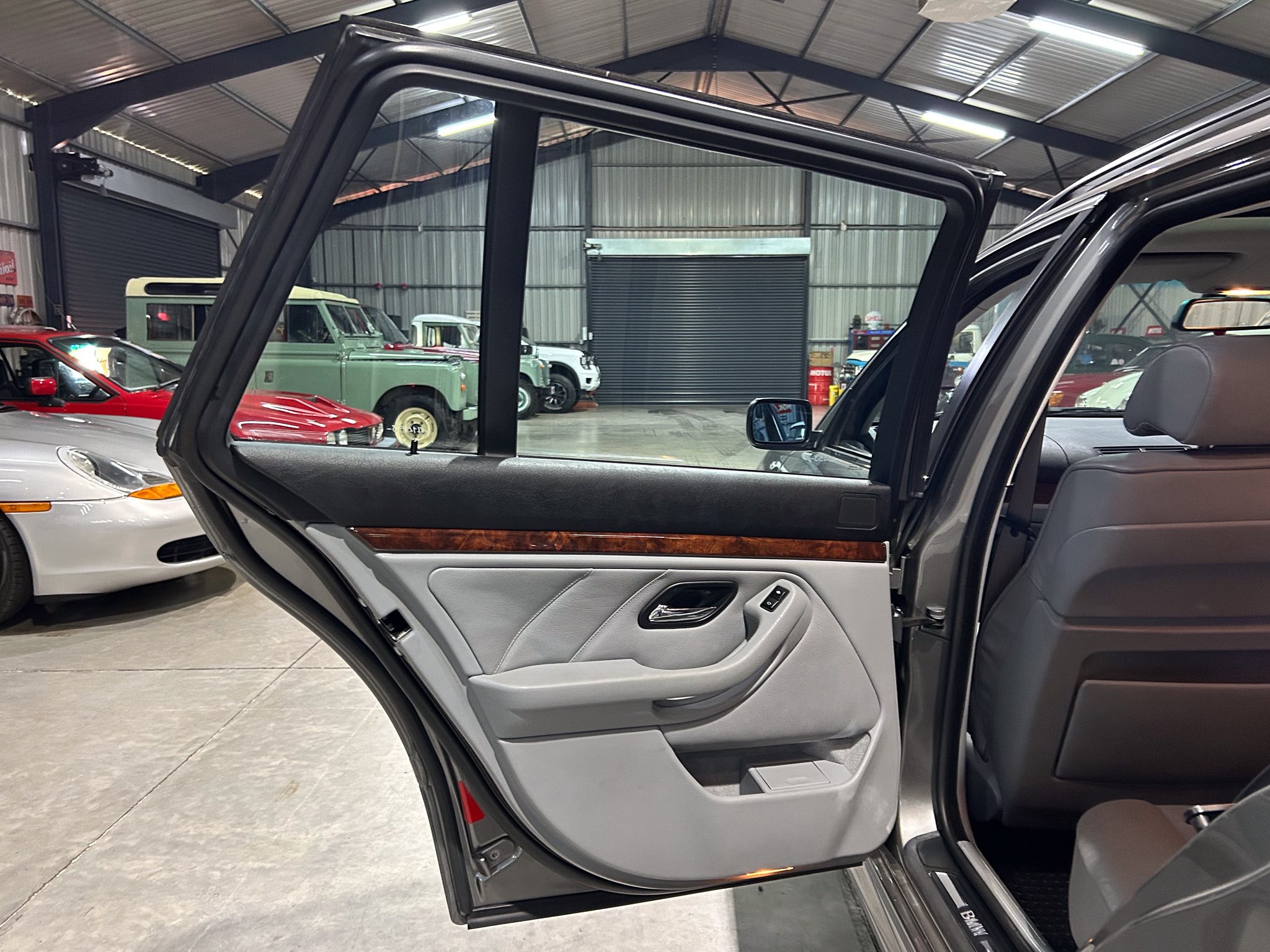 Open rear passenger door of a gray SUV in a showroom, with classic cars visible in the background and a high-ceiling metal workshop.