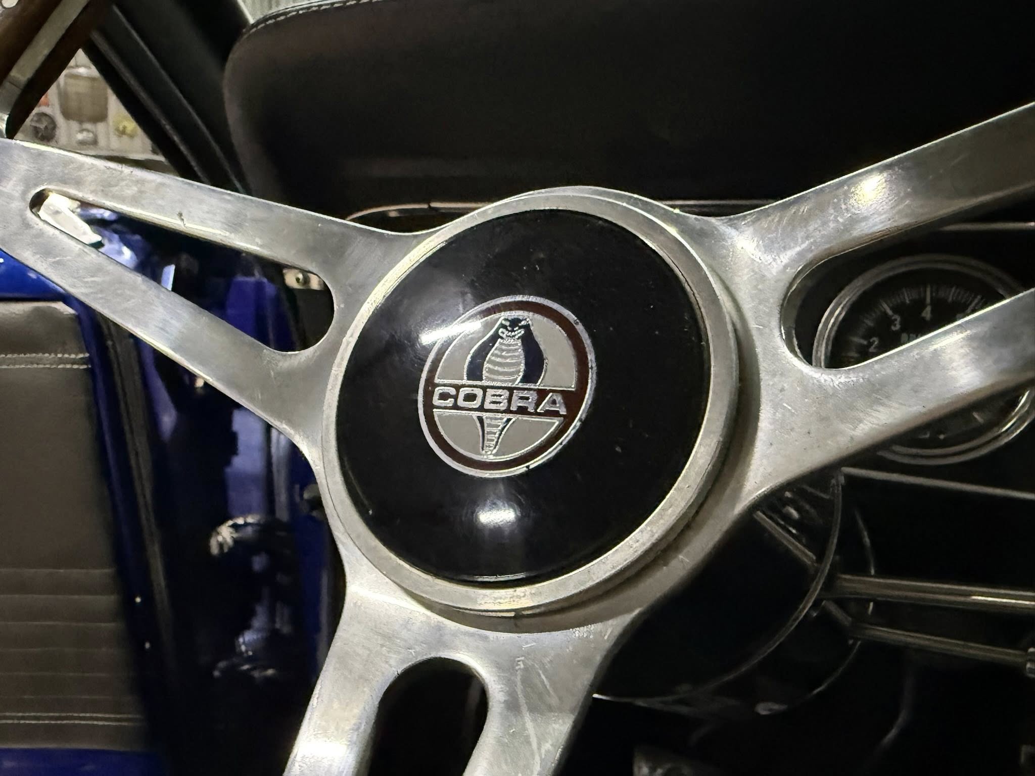 Close-up of a chrome Cobra emblem centered on a multi-spoke metal piece, with part of a gauge visible to the right.