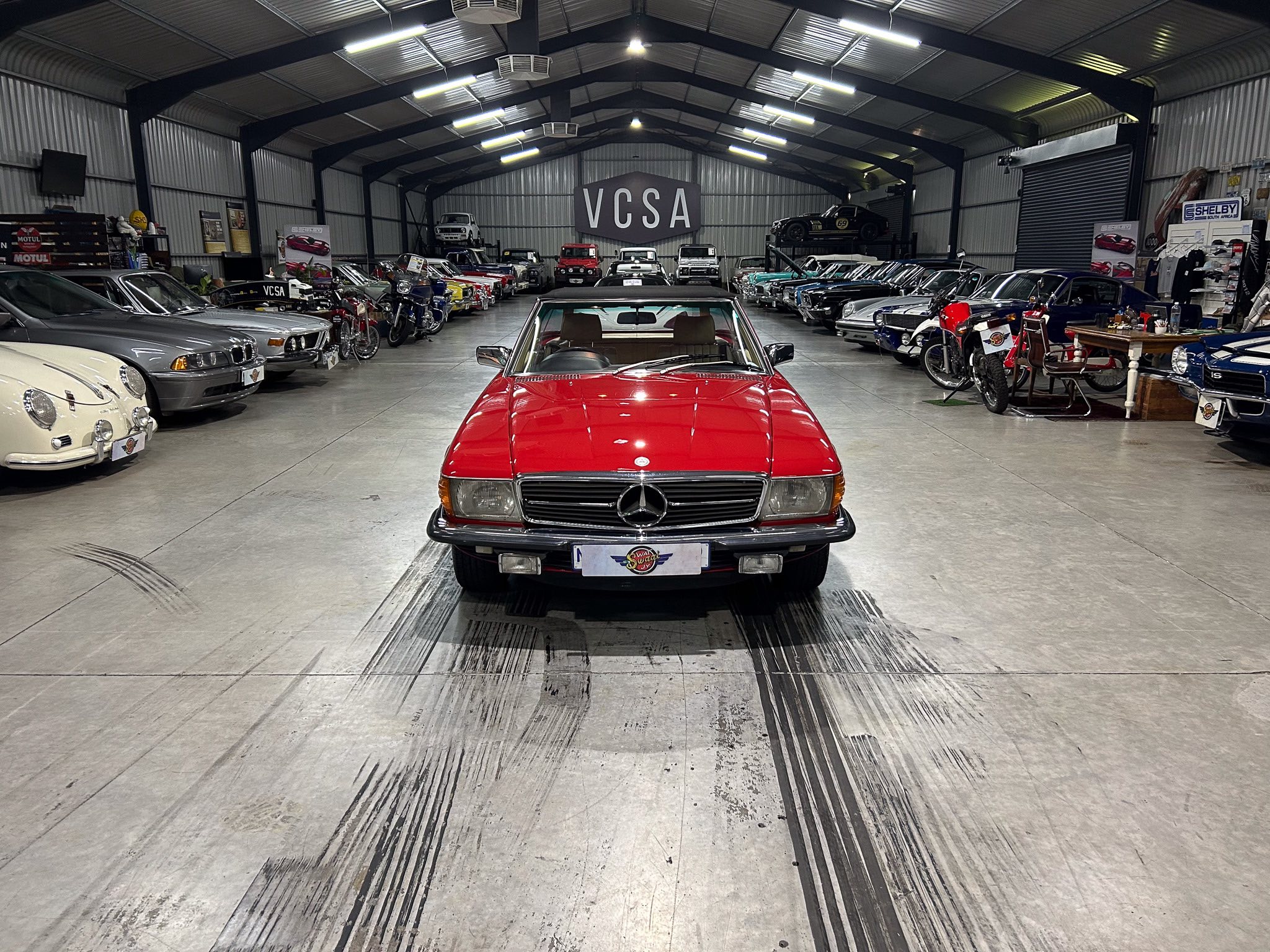 Indoor car showroom filled with classic and vintage cars and motorcycles, centered on a red Mercedes convertible.