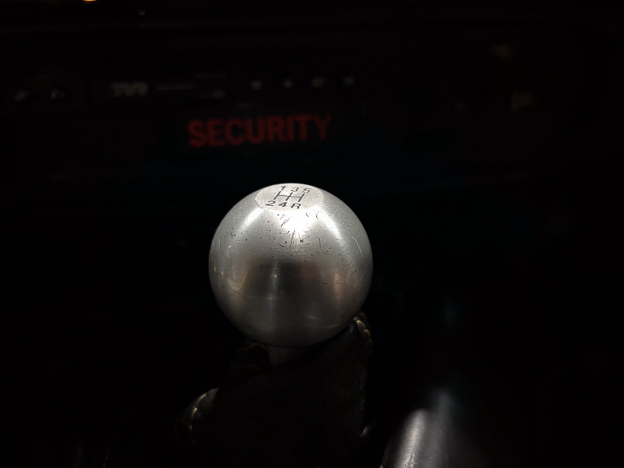 Silver manual gear shift knob showing 1-2-3-4-5 and R in a dark car interior; red 'SECURITY' sign blurred in the background