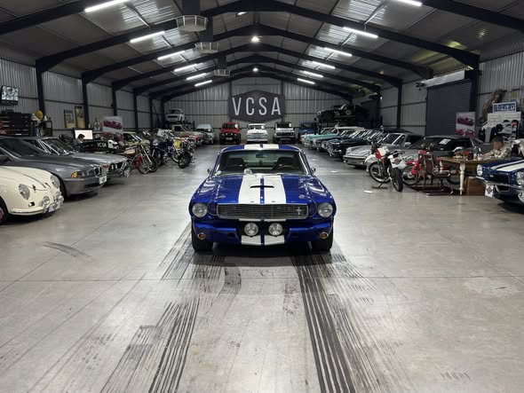 Blue classic car with white racing stripes centered in a showroom filled with vintage cars on both sides.