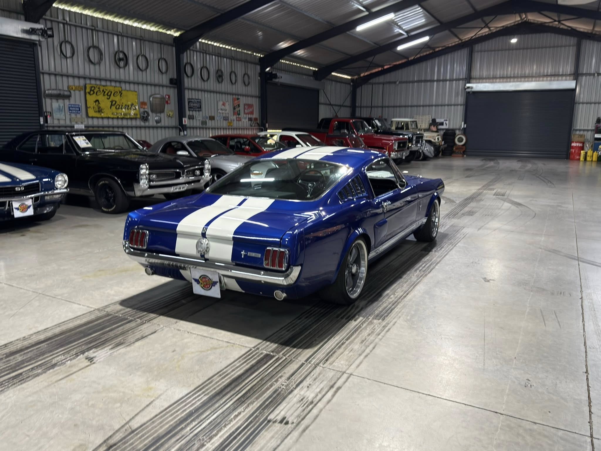 Blue race-striped classic car in the foreground inside a metal-walled garage, with a line of vintage cars along the wall behind it