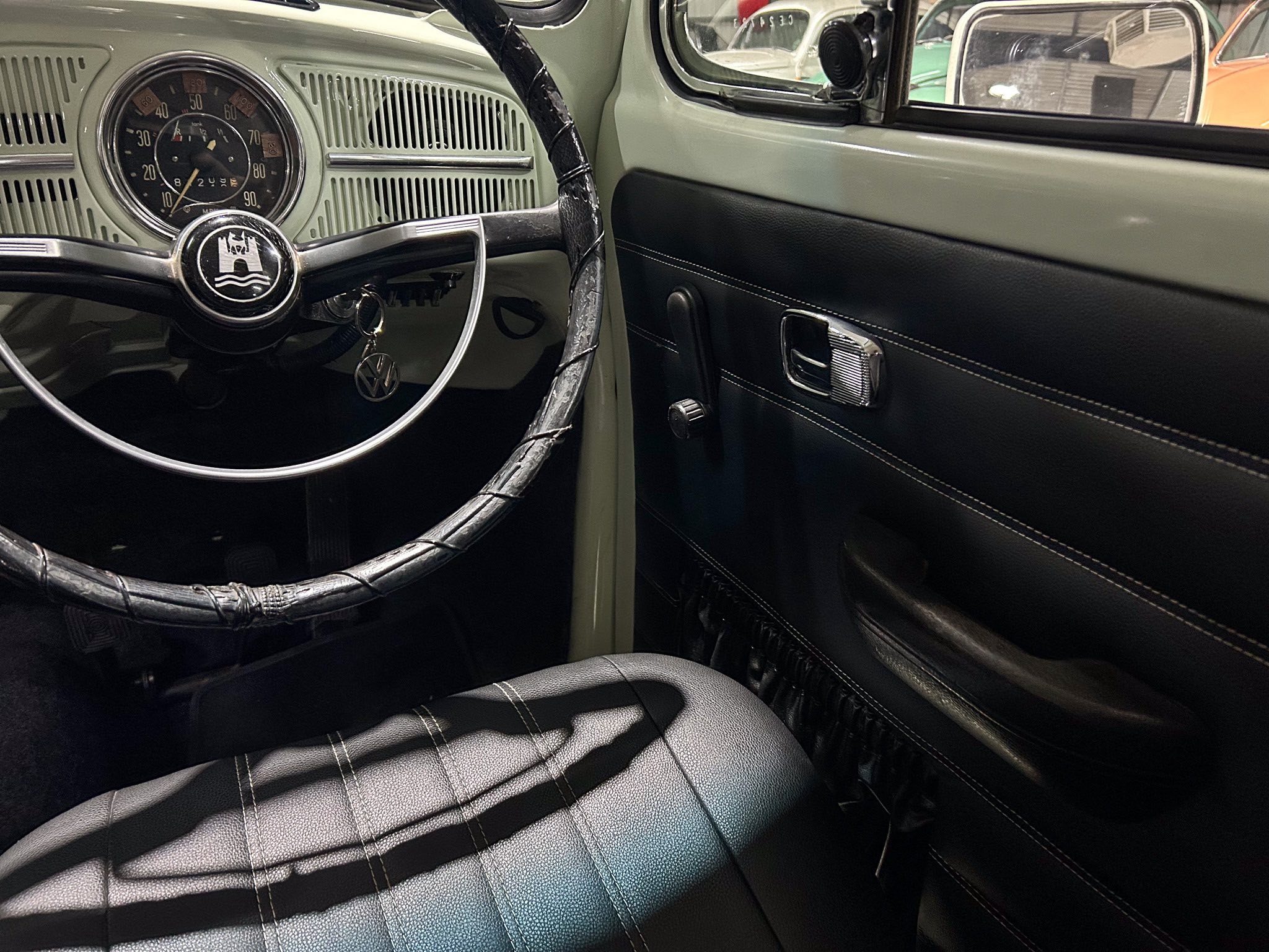 Vintage car interior with a circular gauge cluster, worn steering wheel, and black seats with white stitching on pale dashboard panels