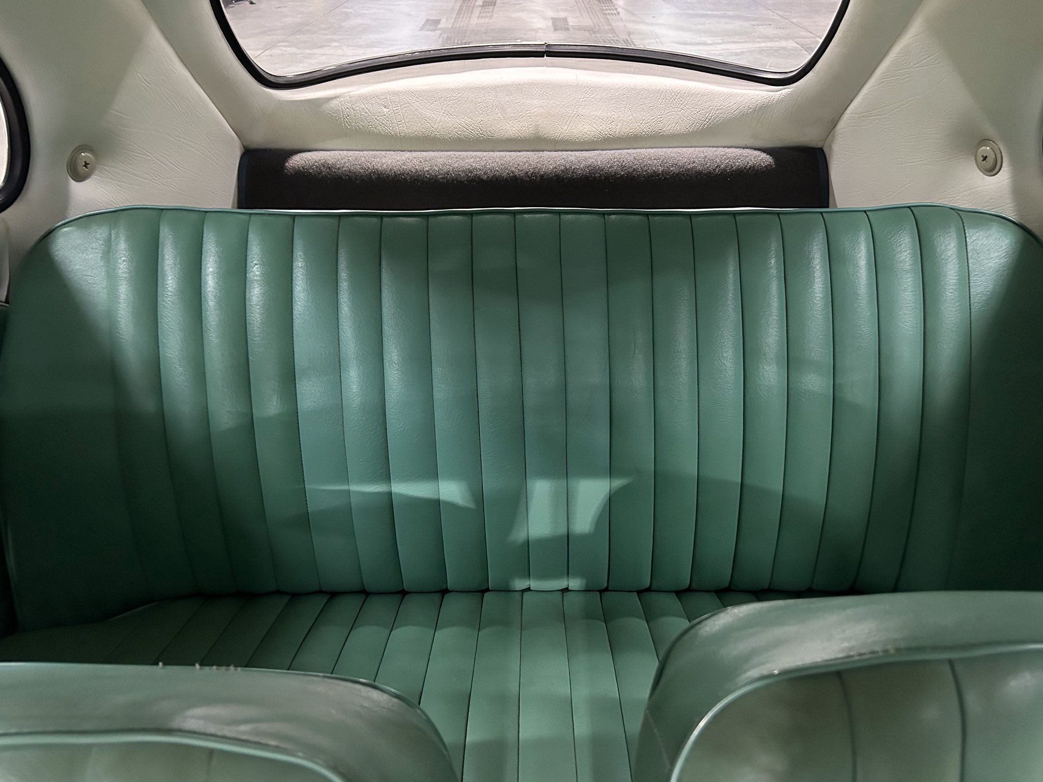 Rear seating area of a vintage car with teal ribbed leather seats and light interior panels, viewed from the backseat toward the rear window.