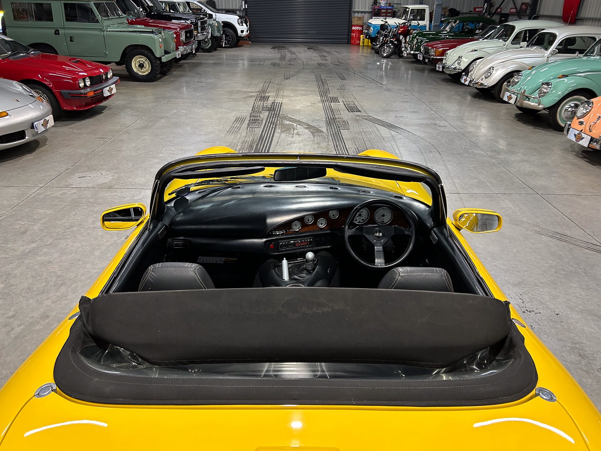 View from inside a yellow convertible facing a spacious showroom filled with classic cars on both sides.