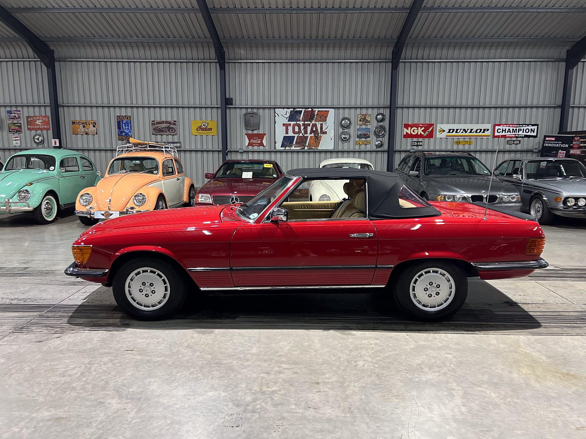 Interior of a vintage car showroom with colorful classic cars lined along a wall of metal panels and retro signs behind them, red convertible in foreground
