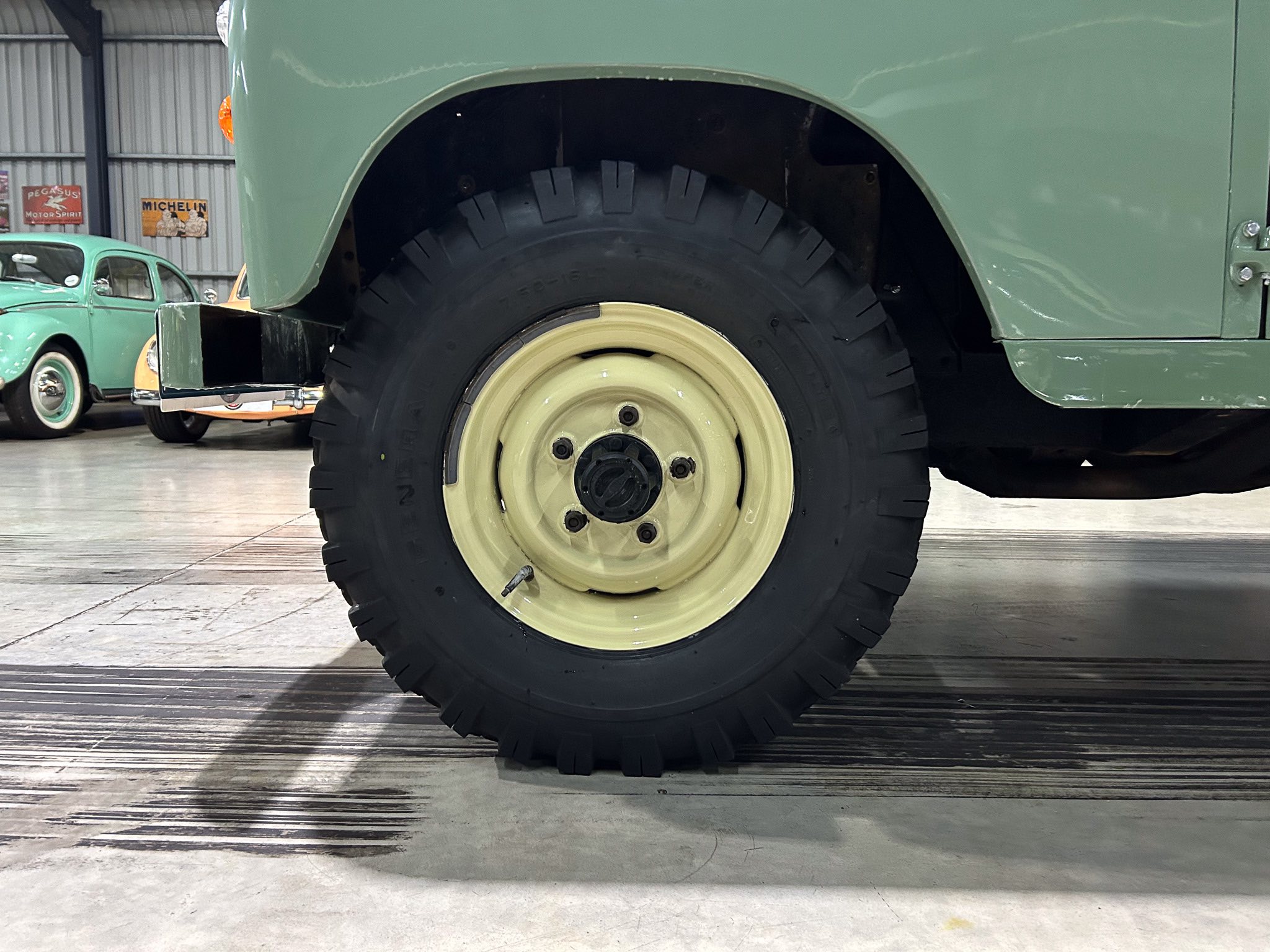 Close-up of a large off-road tire mounted on a cream wheel, attached to a green vintage truck in a museum setting.