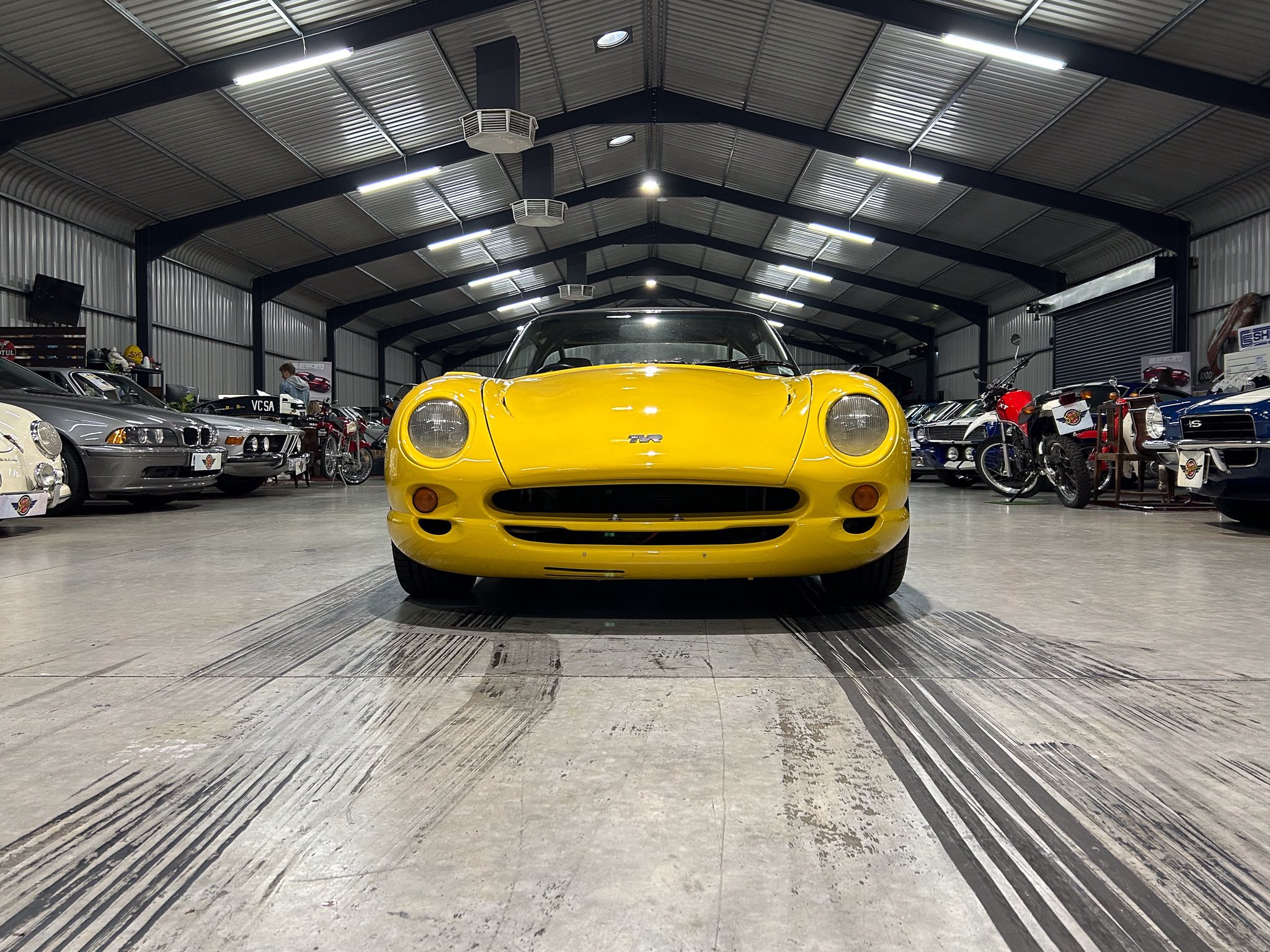 Front view of a bright yellow sports car in a showroom, surrounded by other classic cars and motorcycles under a metal roof.