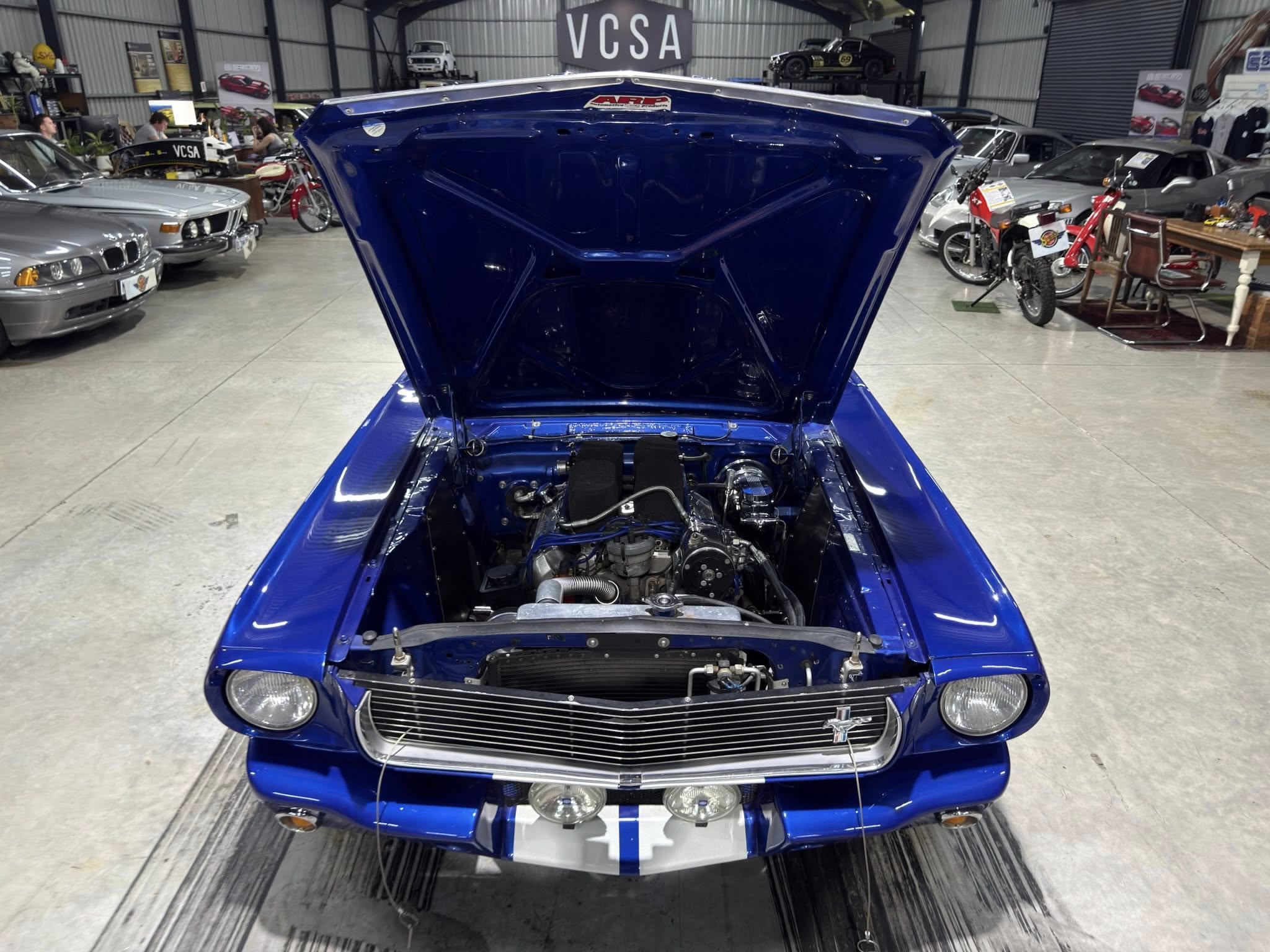 Blue classic car with hood open, showing the engine, inside a showroom with other vehicles and motorcycles in the background.