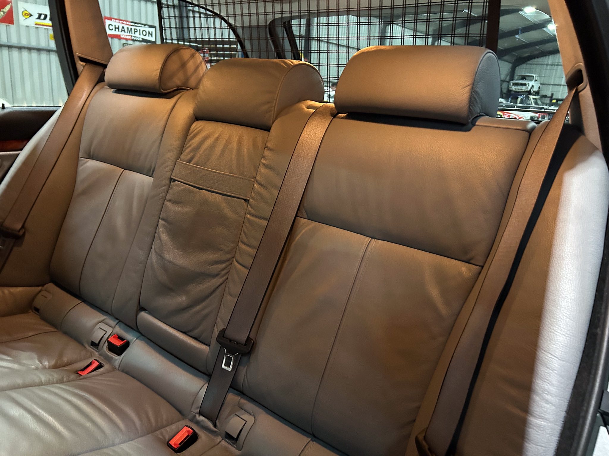 Rear row of a car with tan leather seats, three headrests, and visible seat belts; a metal cargo barrier is behind the seats.