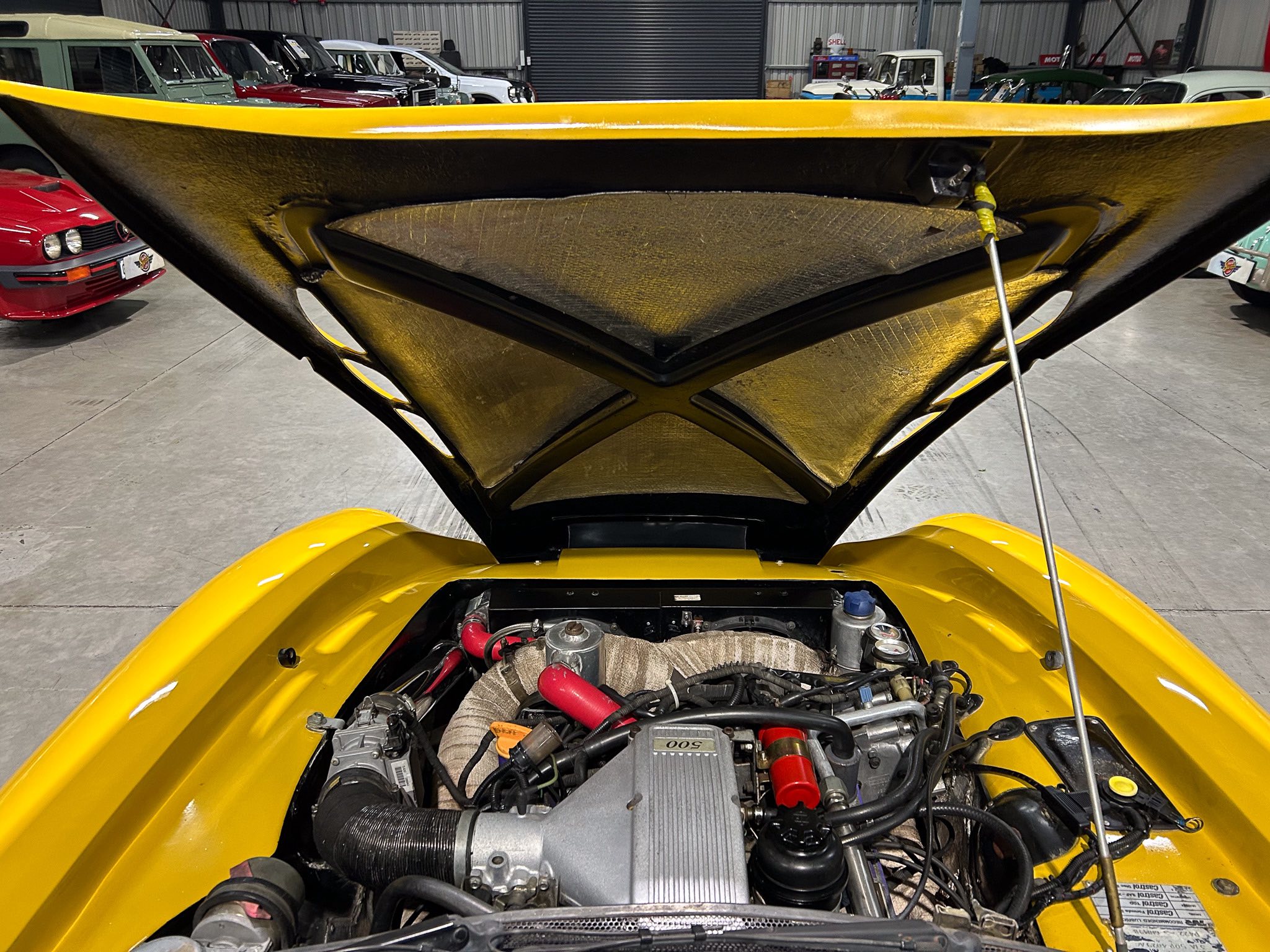 Under-hood view of a bright yellow car, exposing its engine bay with red hoses and metal components in a garage.