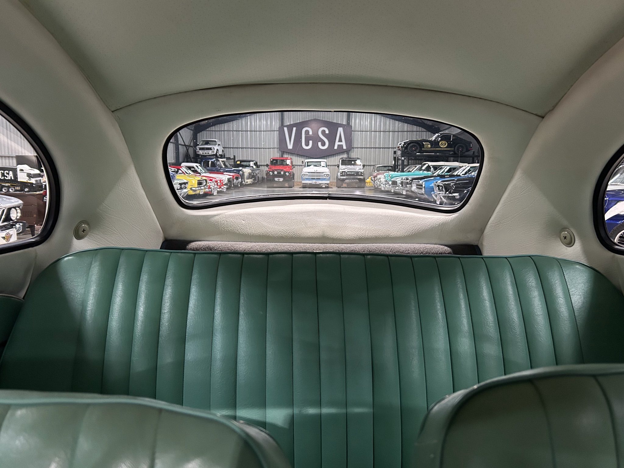 View from the back seat of a vintage car, showing teal leather upholstery and a showroom full of classic cars through the rear window.