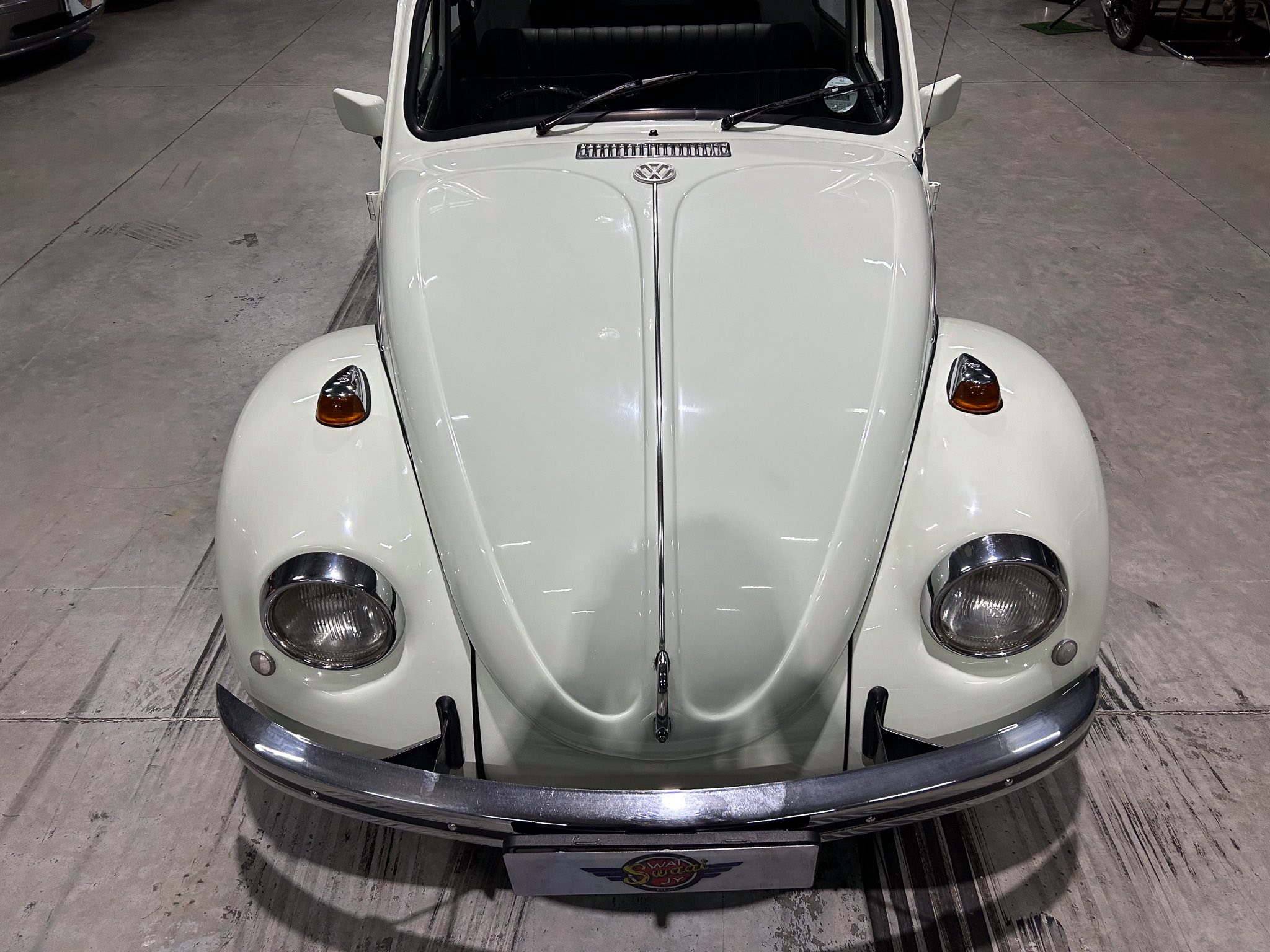 Front view of a pale green vintage Volkswagen Beetle in a showroom, showing its rounded fenders, circular headlights, and chrome bumper from above.