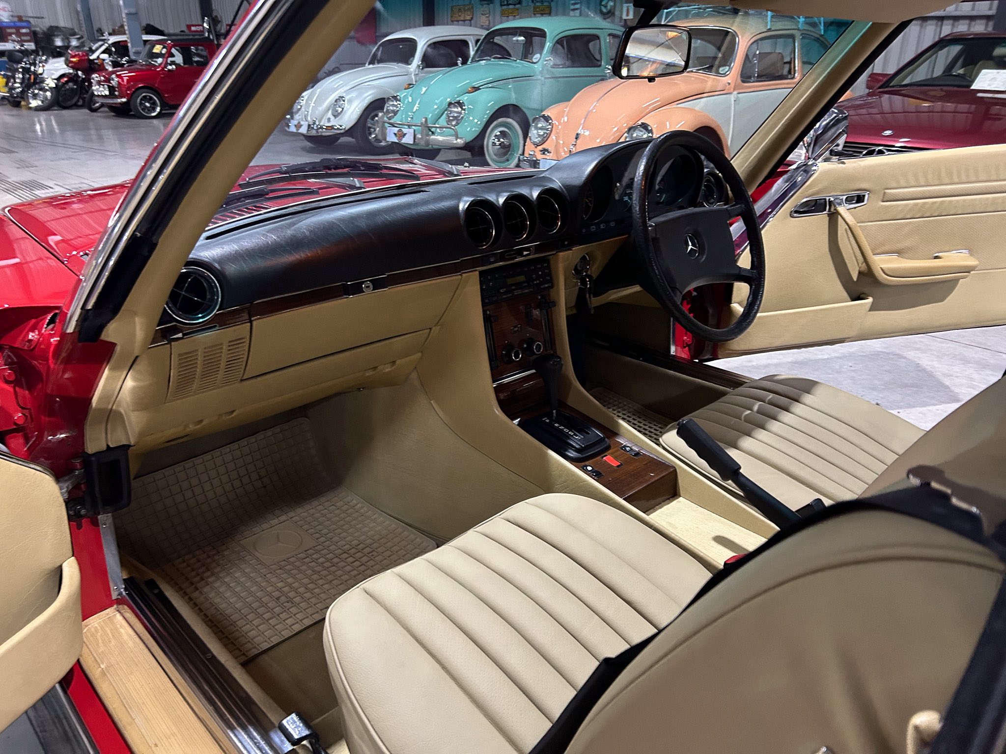 Interior view of a vintage red car with tan leather seats, wood trim, and classic dashboard controls inside a showroom. A row of pastel Volkswagen Beetles is visible outside through the windshield.