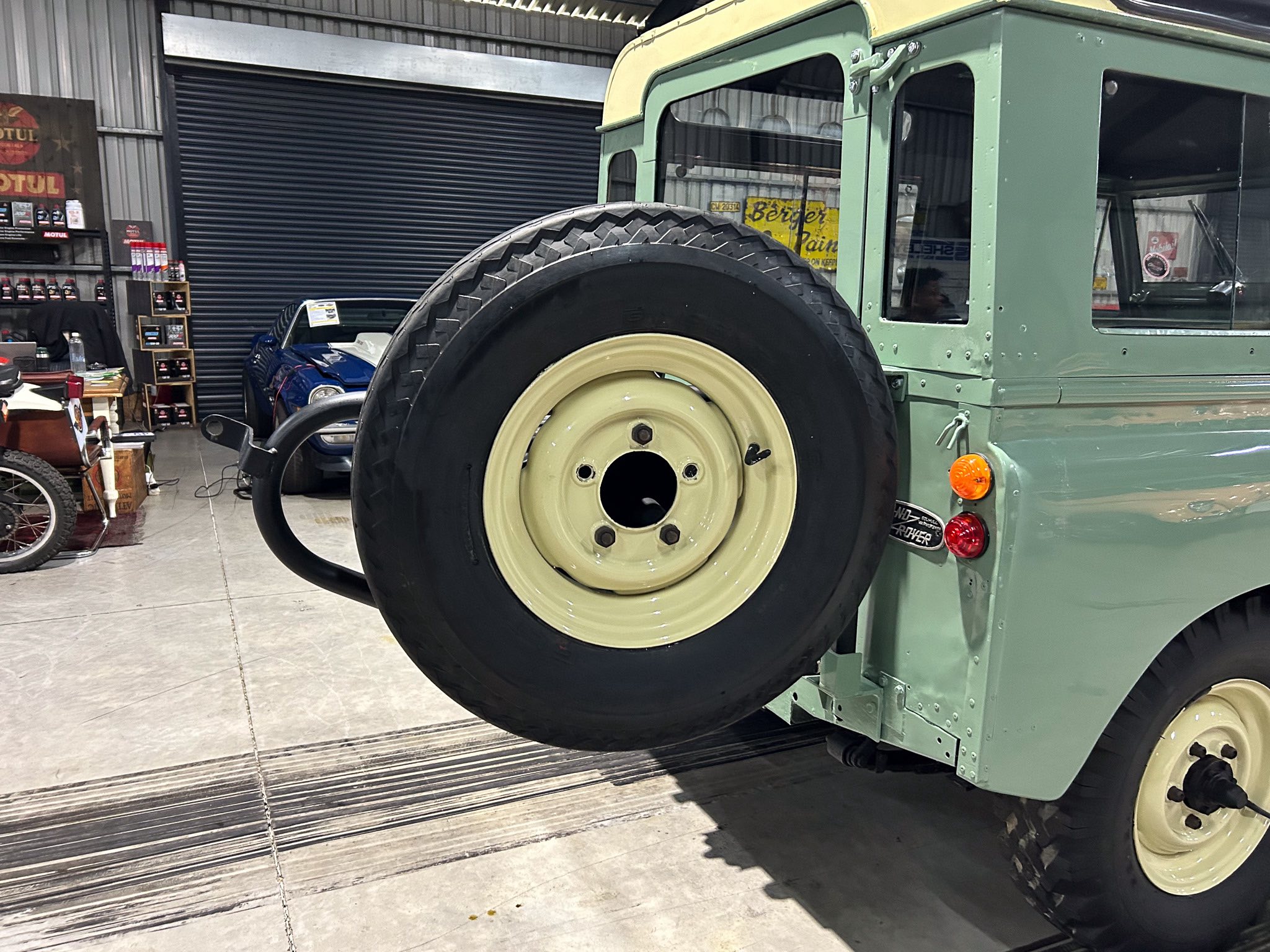 Side view of a vintage green military jeep with a large spare tire mounted on the left, inside a workshop with shelves of oil and a blue car in the background.