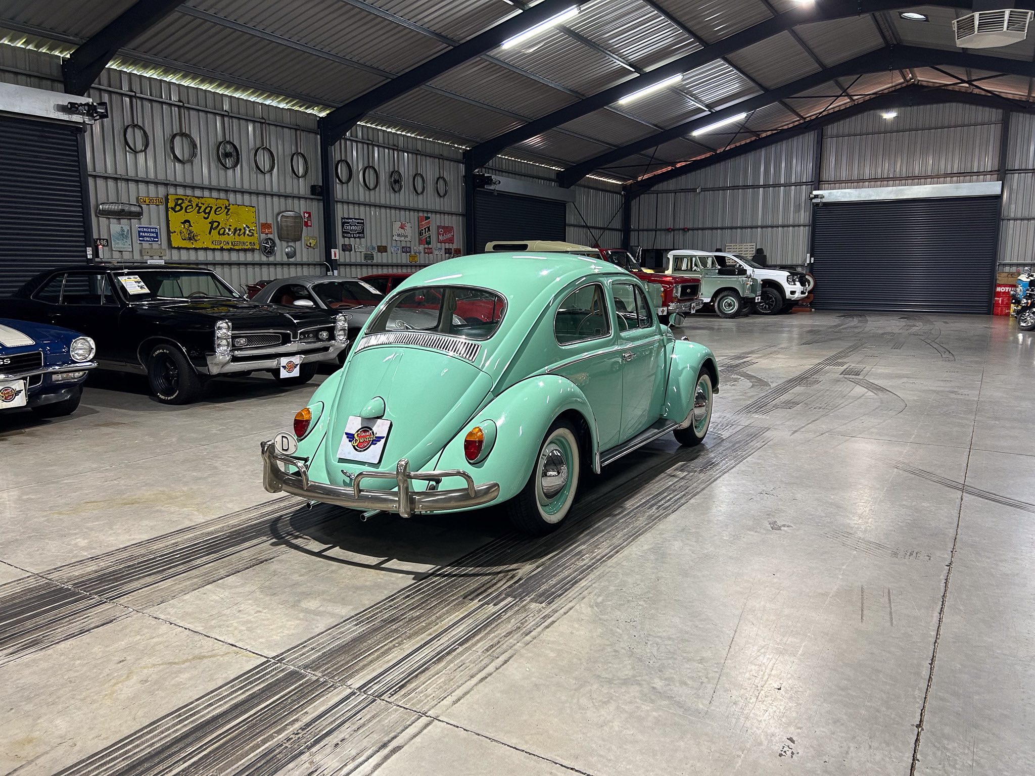 Mint green vintage Volkswagen Beetle in a warehouse showroom with other classic cars and wall signs behind it.