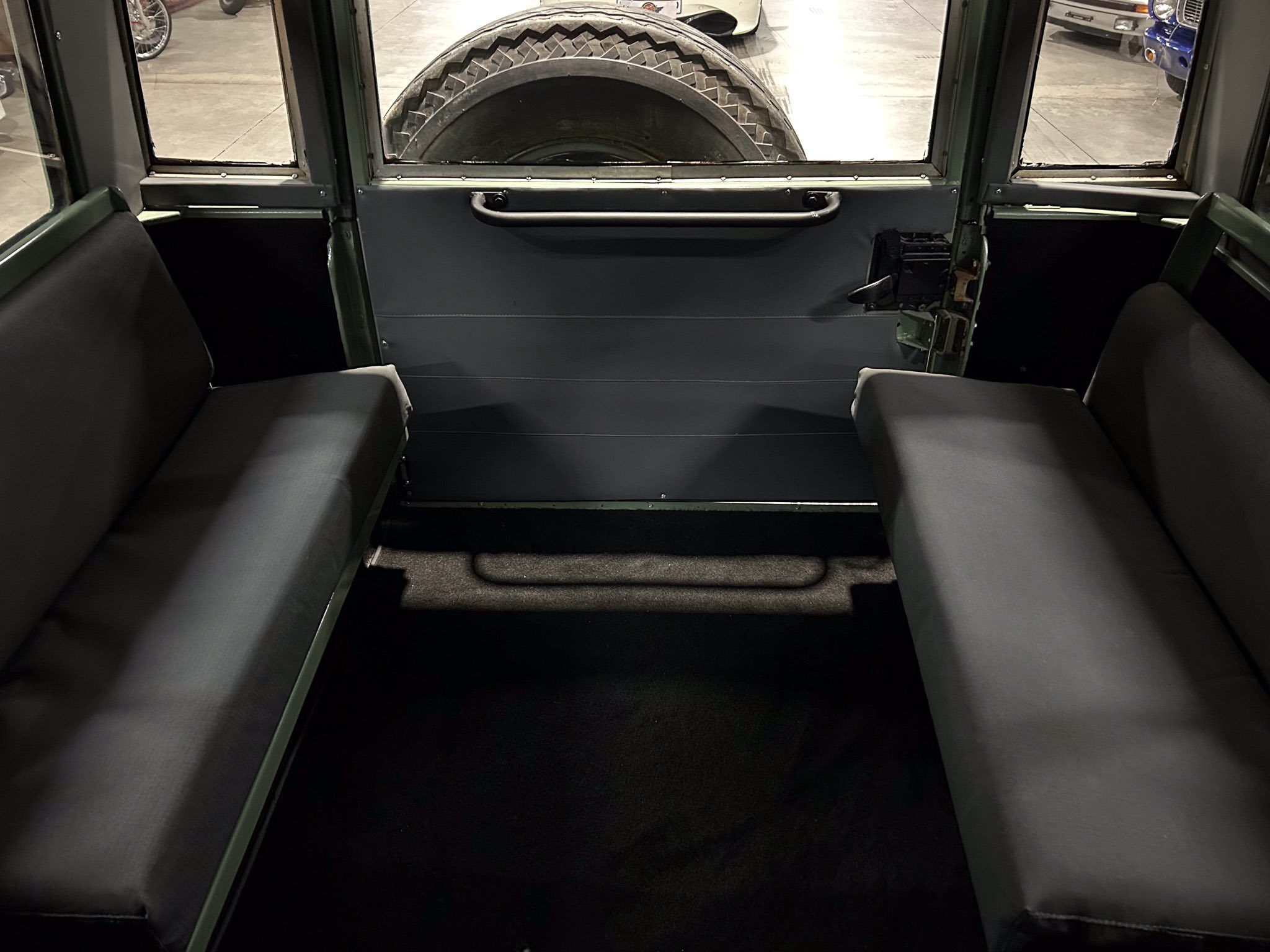 Interior of a vehicle cabin with two long bench seats along the sides and a large spare tire visible through the rear hatch window, viewed from the back of the cabin.