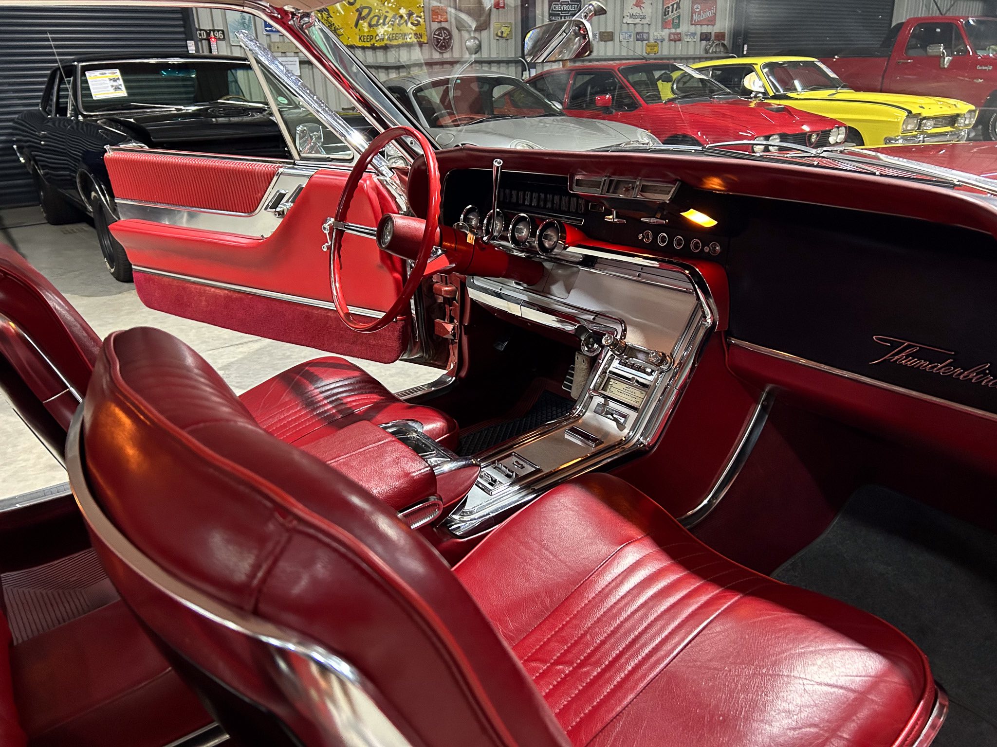 Interior view of a red vintage car with red leather seats, a red steering wheel, and chrome dashboard accents in a showroom filled with other classic cars.