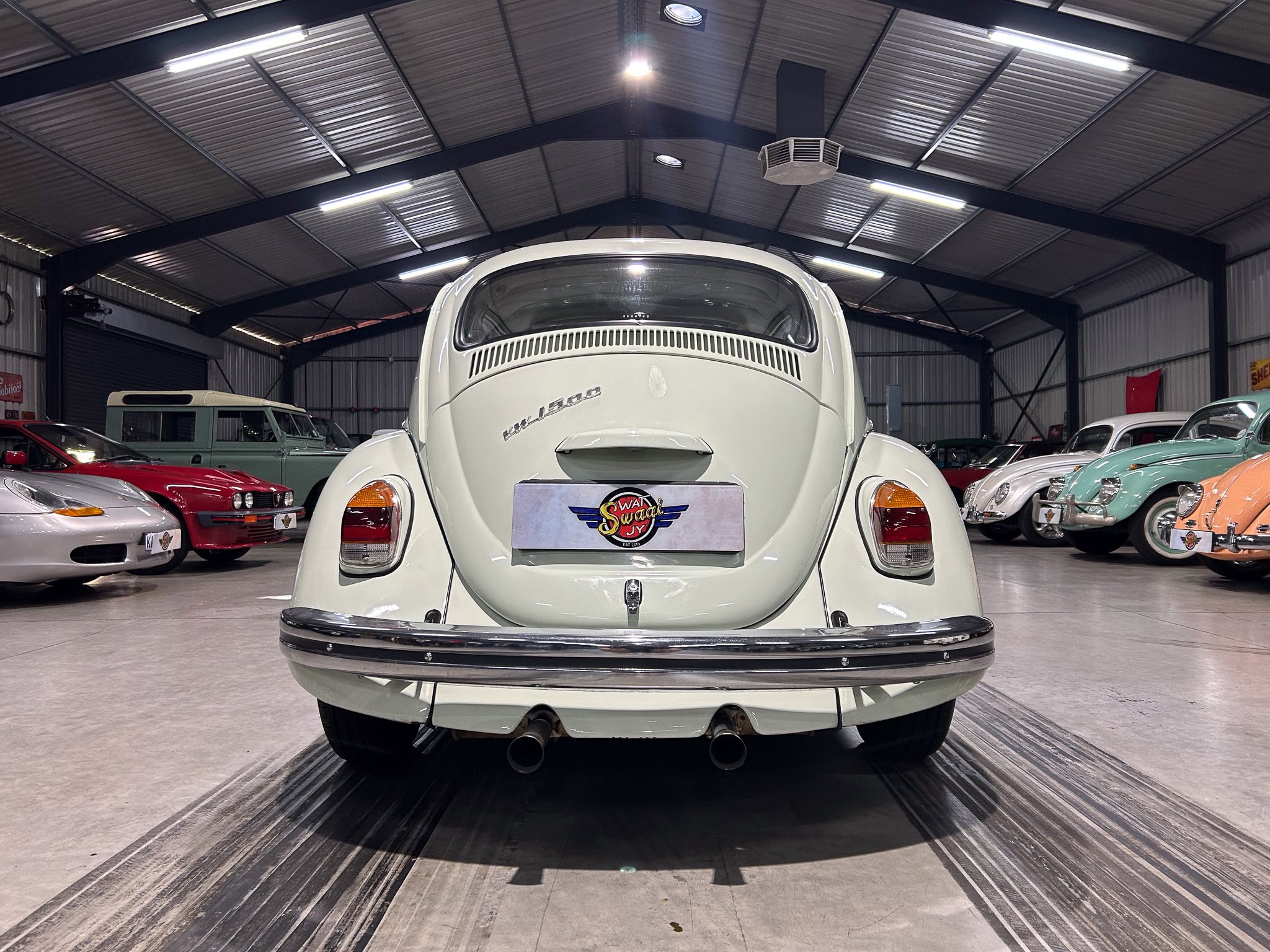 Rear view of a white vintage Volkswagen Beetle in a large showroom, surrounded by other classic cars.