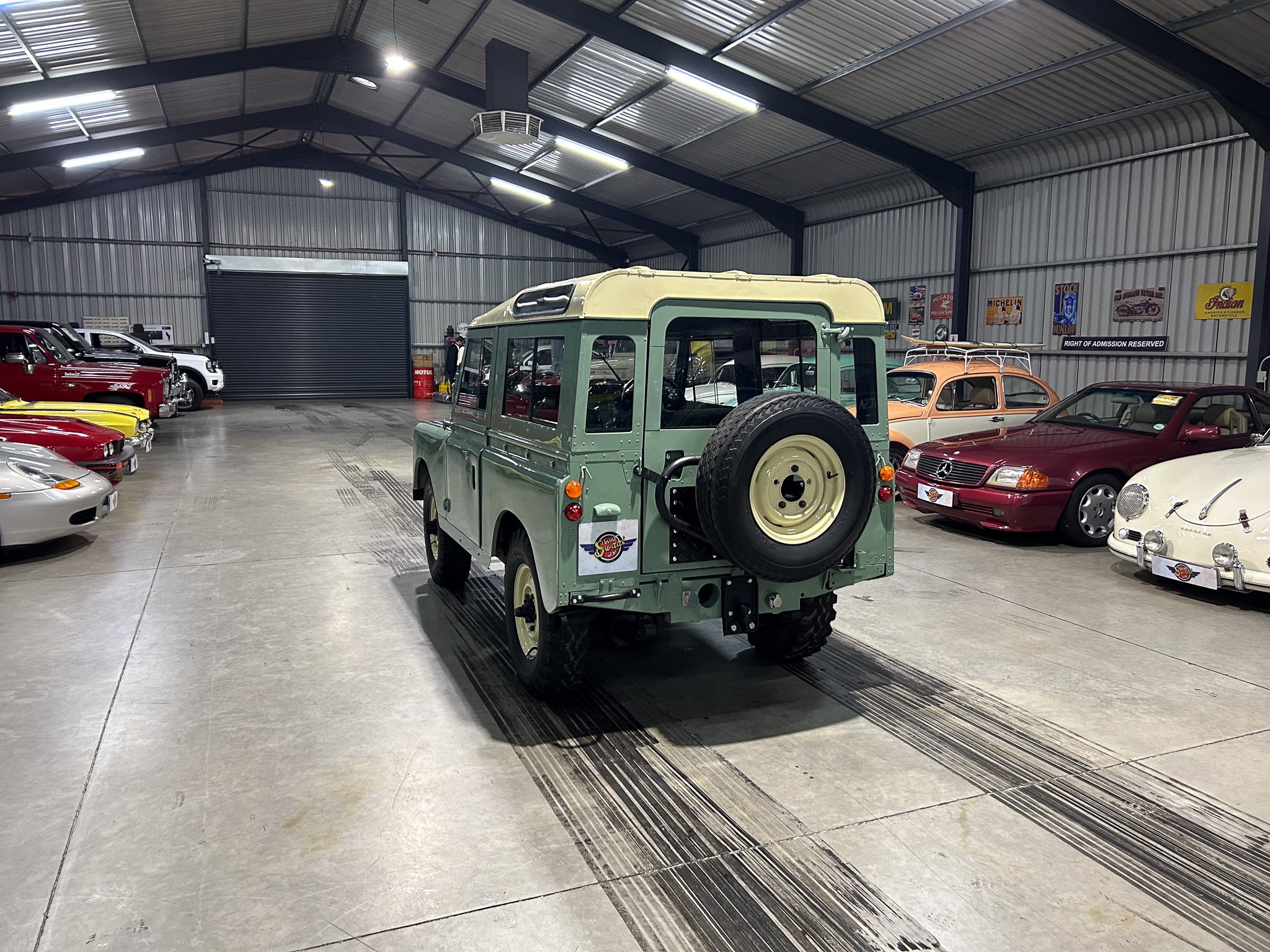 Interior of a large metal warehouse filled with vintage cars, centered on a green vintage SUV with a spare tire on the back and banners on the wall.